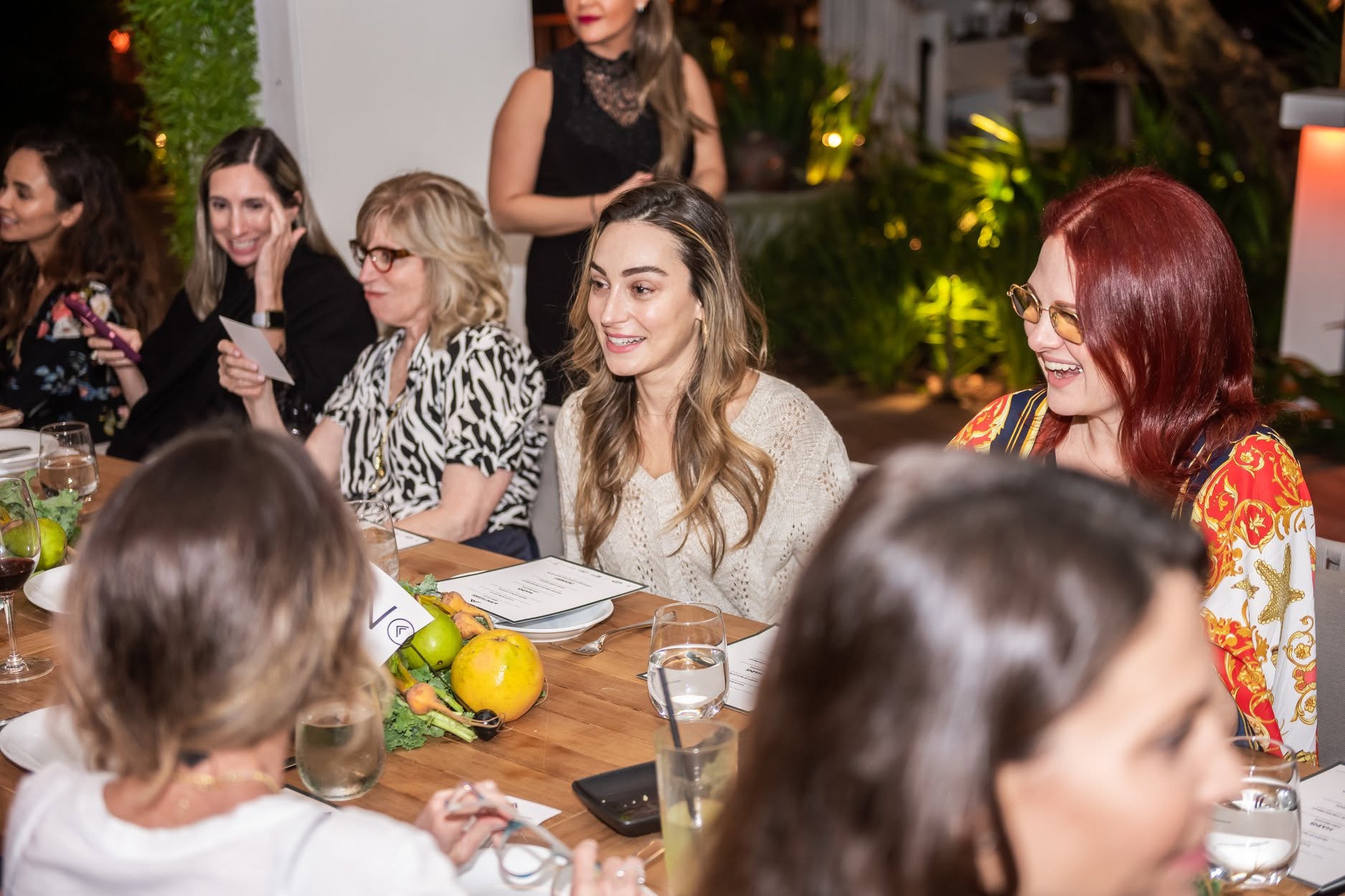 Group of women sitting at a table outdoors, smiling and talking, with drinks and fruits on the table.