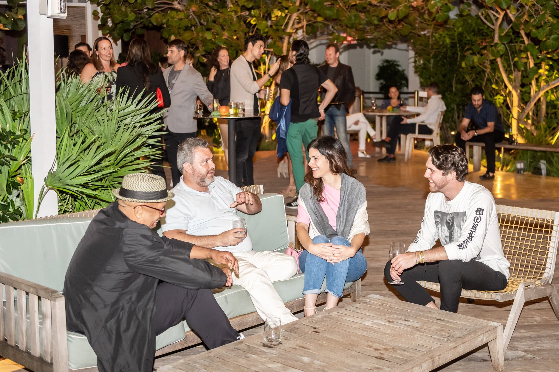 Group of four people sitting and talking outdoors, with others in the background, trees, and tables, during evening.