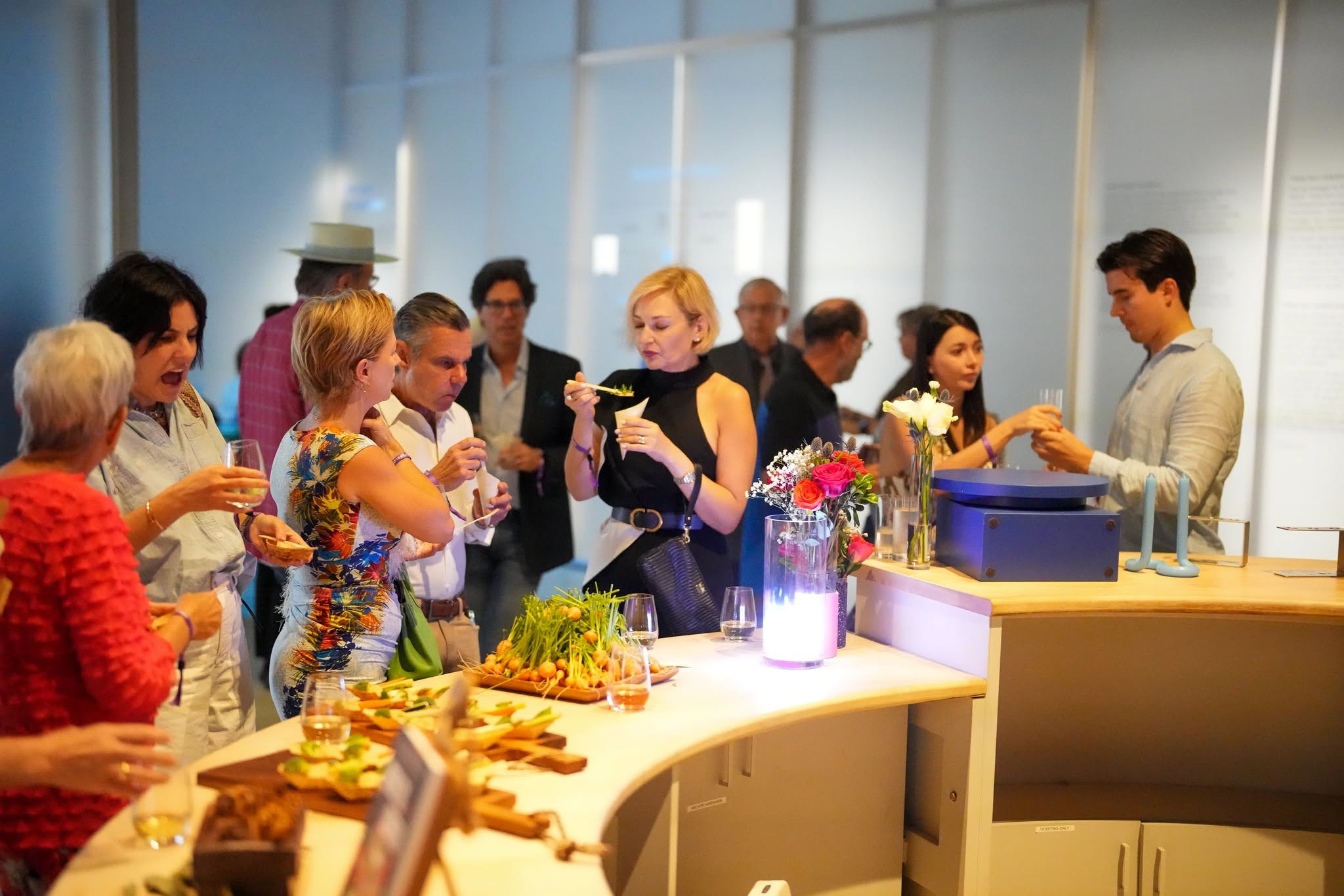 People gather around a buffet table with food and drinks in a bright, modern room with large windows.