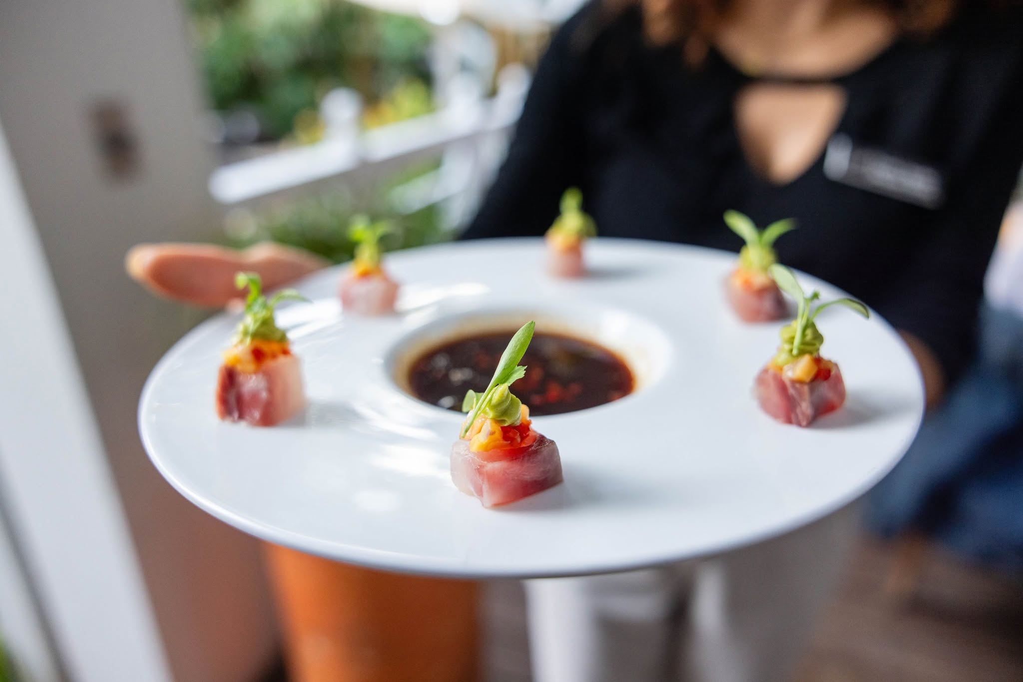 Person holding a white plate with six small appetizers garnished with green herbs, arranged in a circle, with a dark dipping sauce in the center.