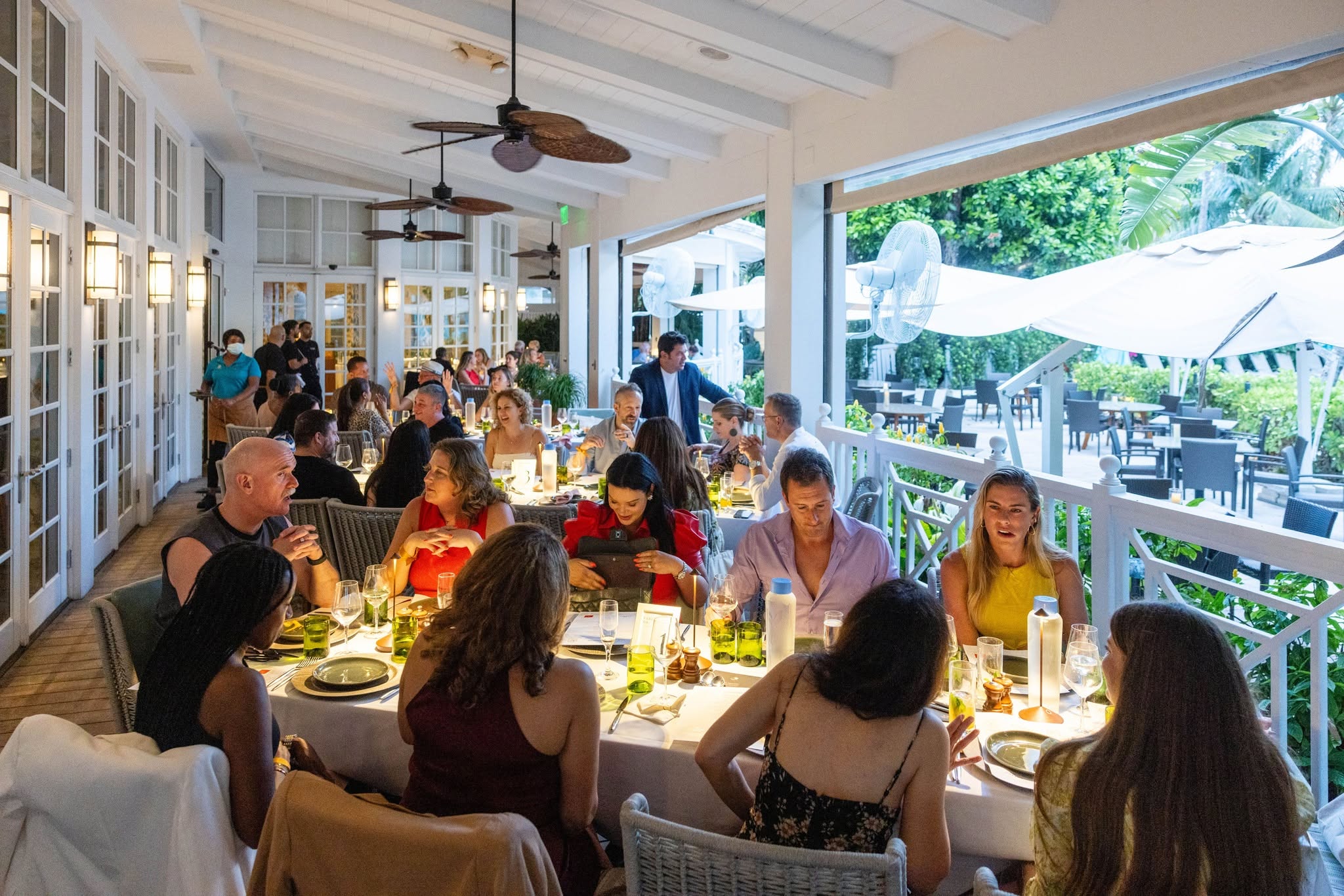 People dining at tables in a bright, open restaurant with large windows and outdoor patio, ceiling fans overhead.