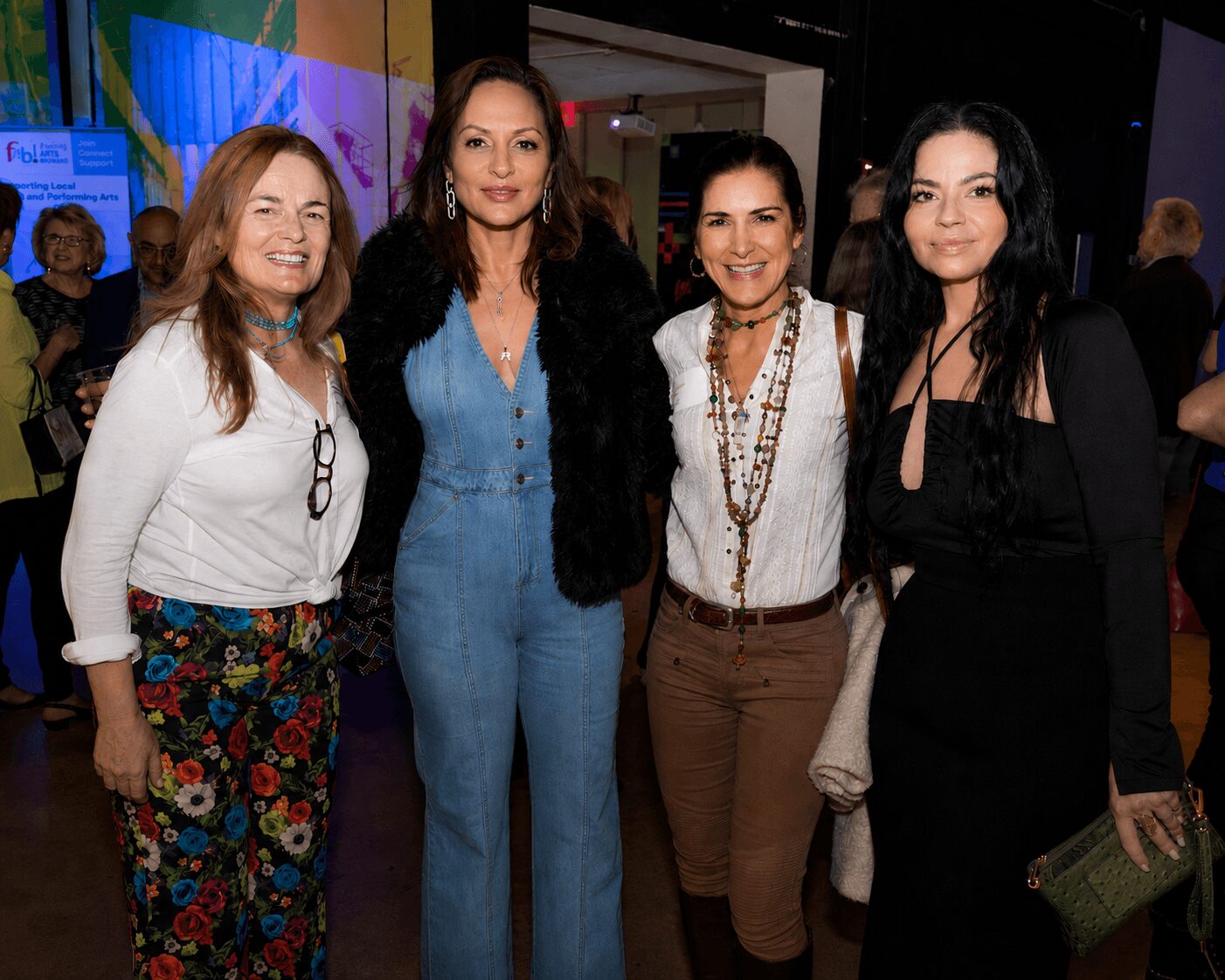 Four women standing together indoors, smiling, with colorful background and people in the distance.