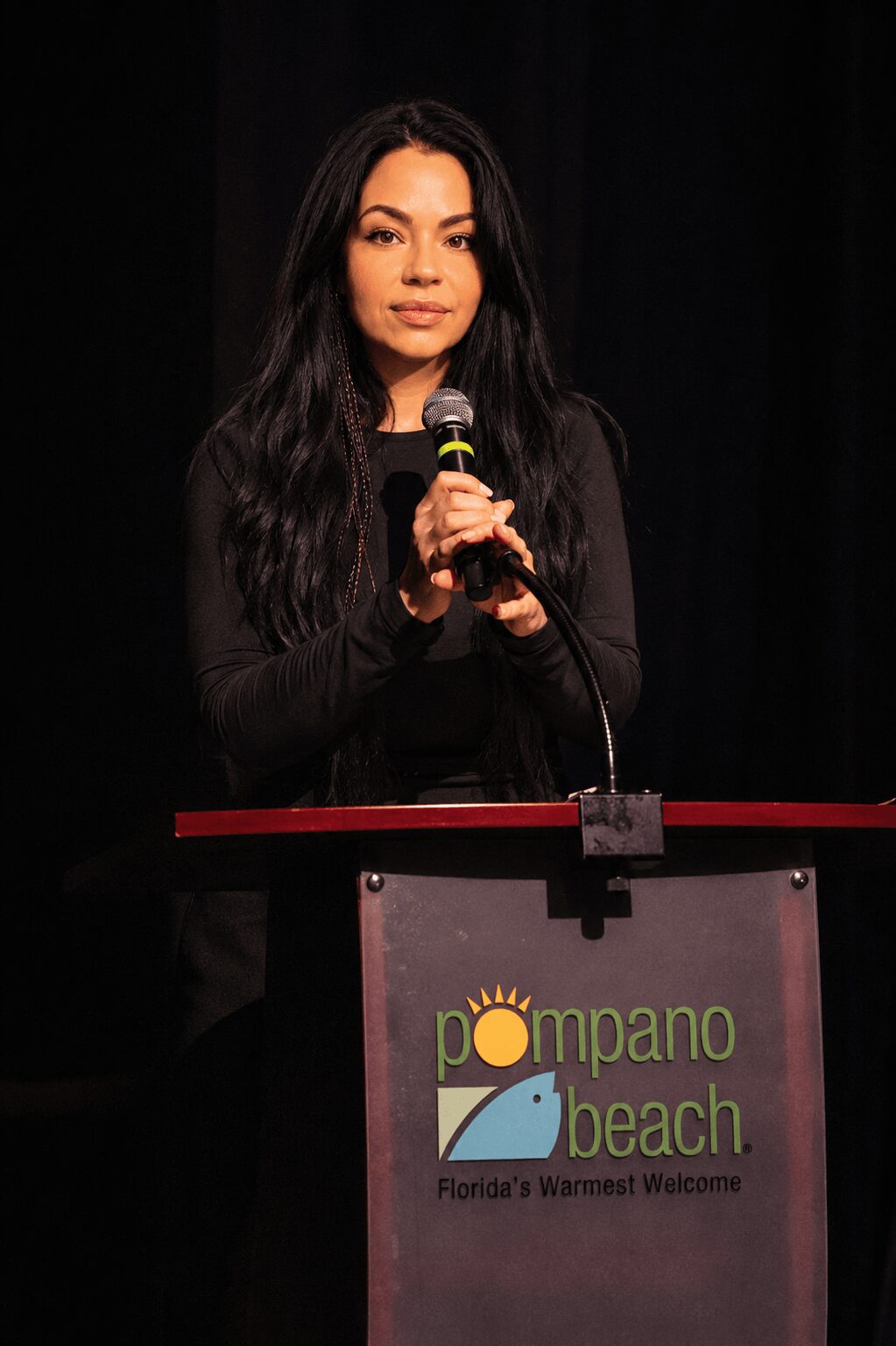 Young woman with long dark hair holding a microphone, standing behind a podium with 'Pompano Beach' logo, black background.