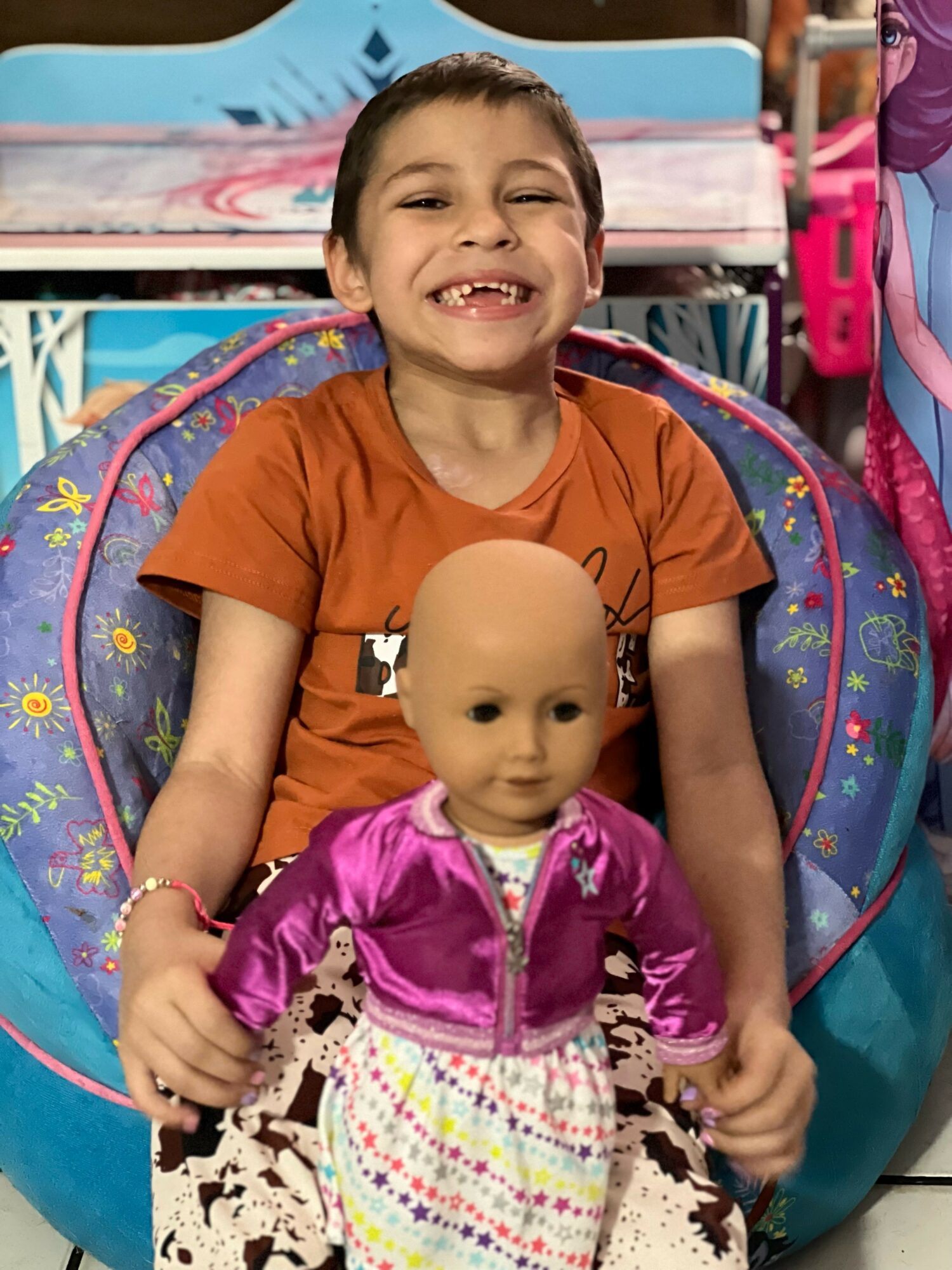 Smiling child sitting in a colorful chair holding a doll, with a background of toys and a pink backpack.