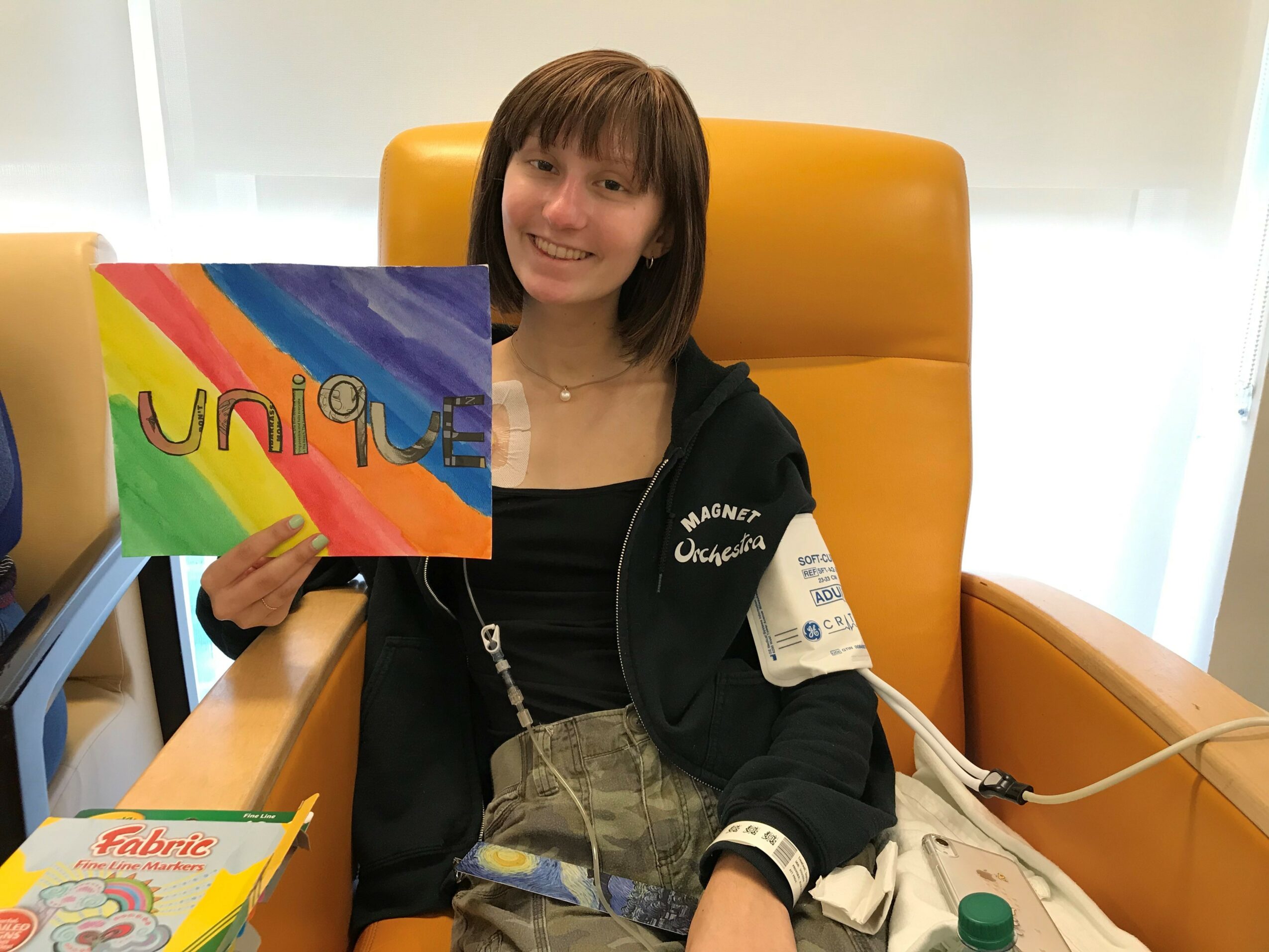 Young girl with brown hair smiling, holding a colorful rainbow sign with the word 'unique', sitting in a yellow chair.