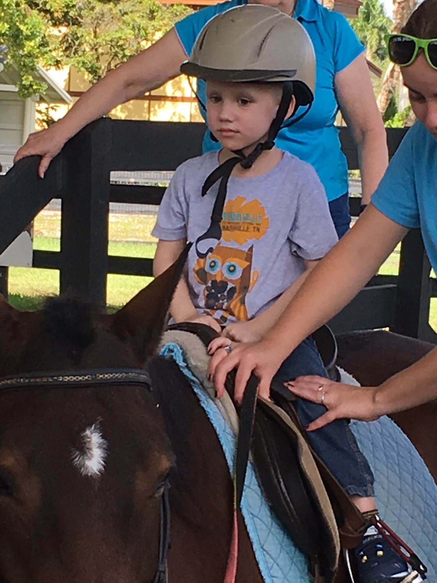 Child wearing a helmet and a gray t-shirt with an owl graphic, sitting on a horse, with two people assisting outdoors.