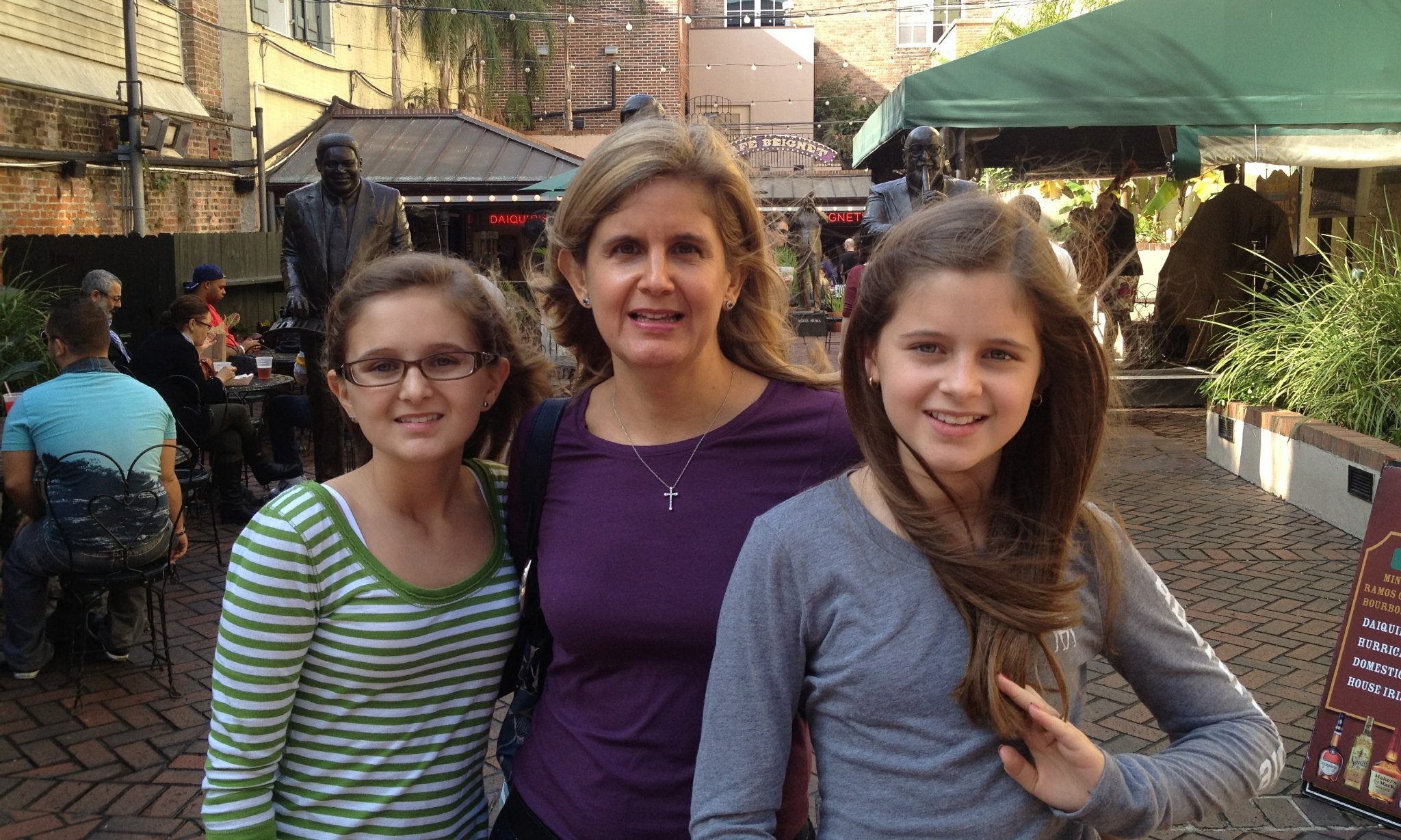 Three women smiling outdoors in a market or street setting, with people and tents in the background.