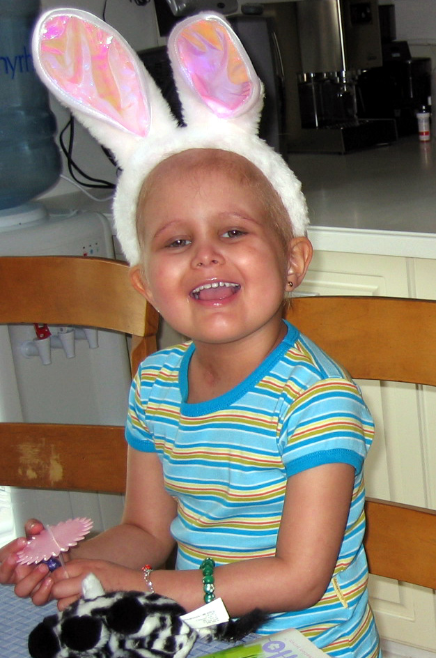 Child wearing bunny ears headband, smiling, sitting at a table with a pink toy, in a kitchen setting.