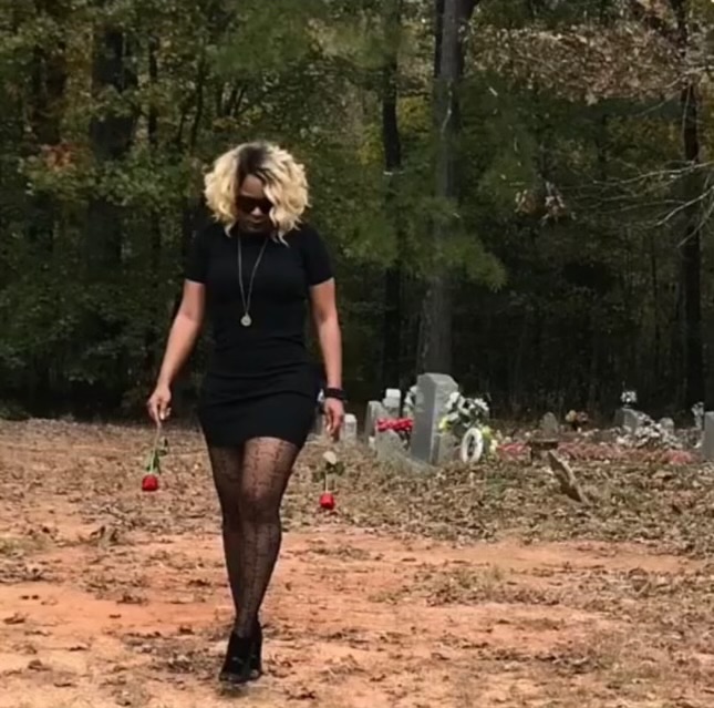 Woman walking on dirt path in a wooded area, wearing black dress, patterned tights, sunglasses, and a necklace.