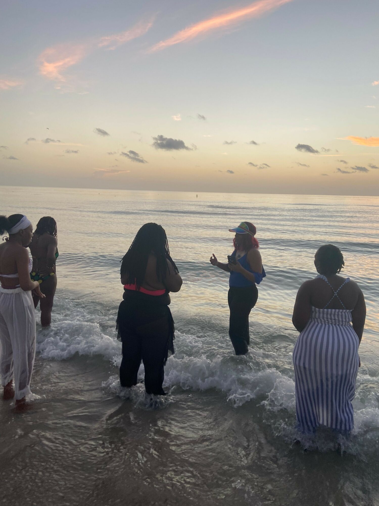 Three people standing in shallow water at the beach during sunset, with the sky showing orange and pink hues.