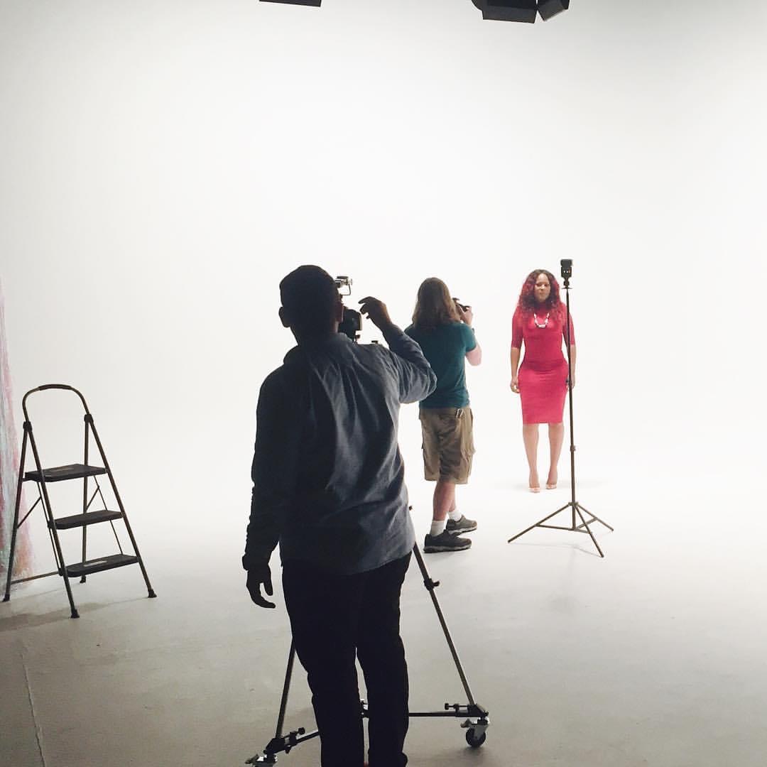 Person filming a woman in a pink dress on a white studio background, with a ladder and lighting equipment nearby.