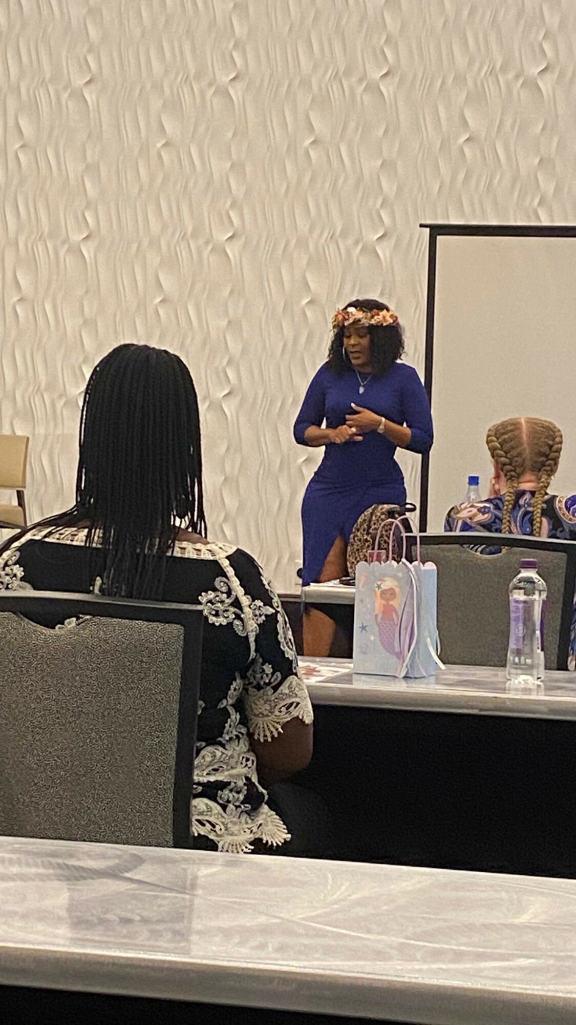 Woman in blue dress standing and speaking to a group in a conference room, with a whiteboard and table in front.