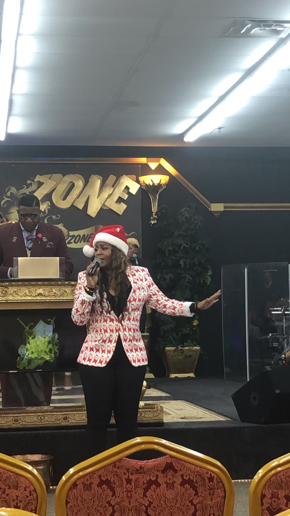 Person speaking into microphone on stage with a Christmas hat, in front of a sign that says 'ZONE'.