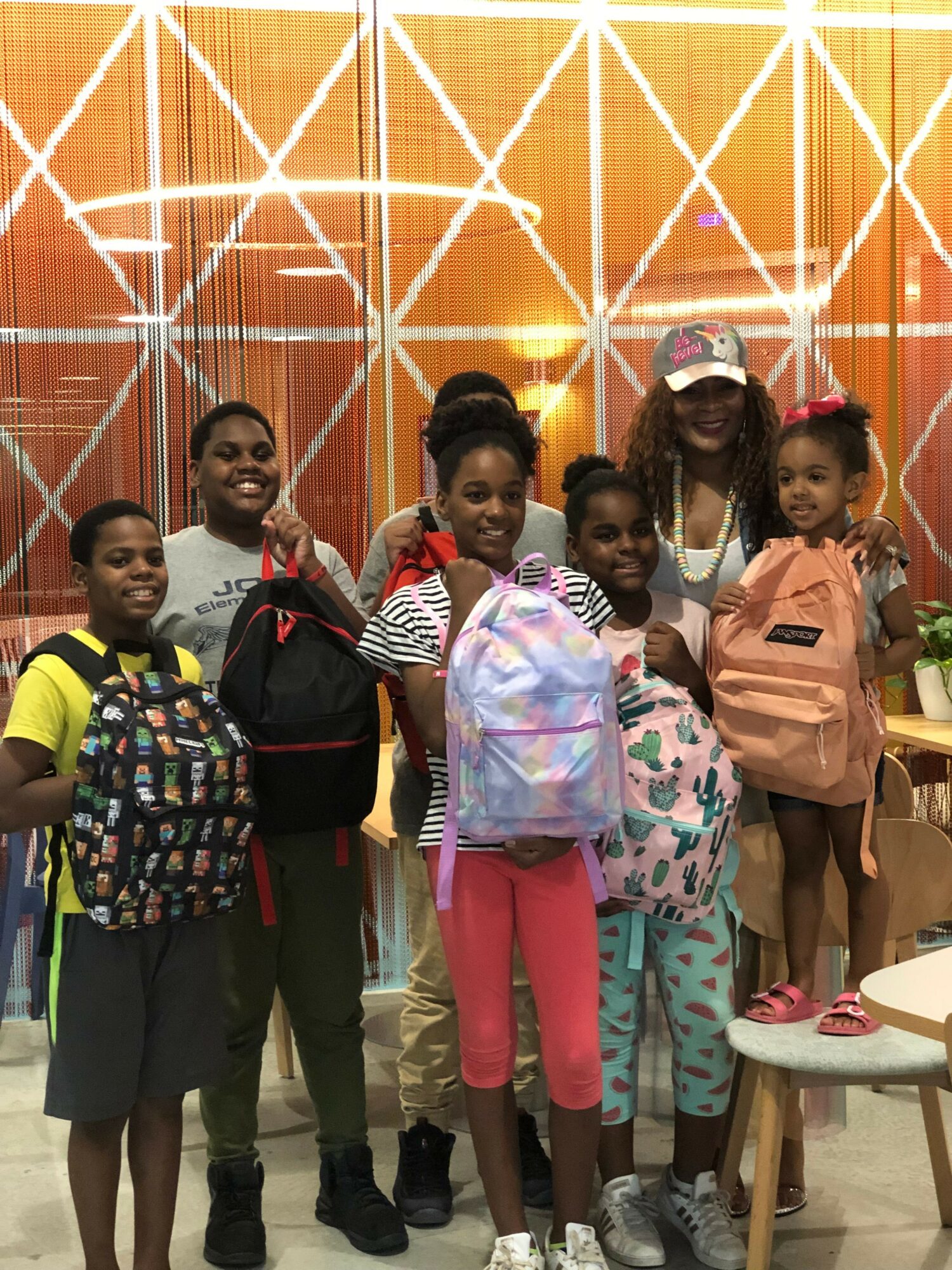 Group of children standing indoors in front of a decorative orange wall, some holding bags, wearing colorful clothing.