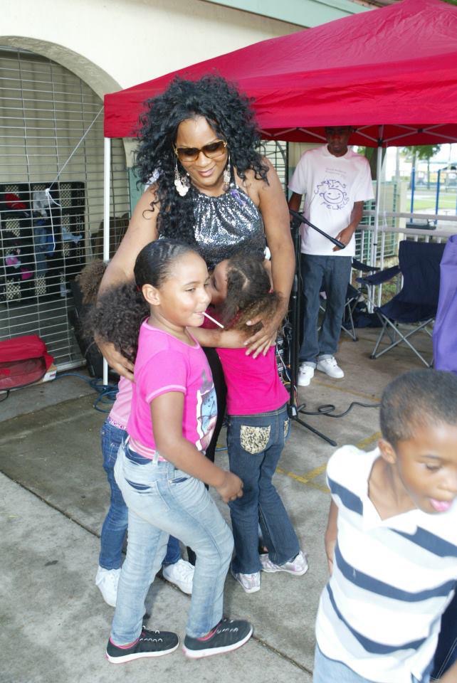 Woman with curly hair and sunglasses standing with children outdoors near a red canopy, some children are smiling and talking.
