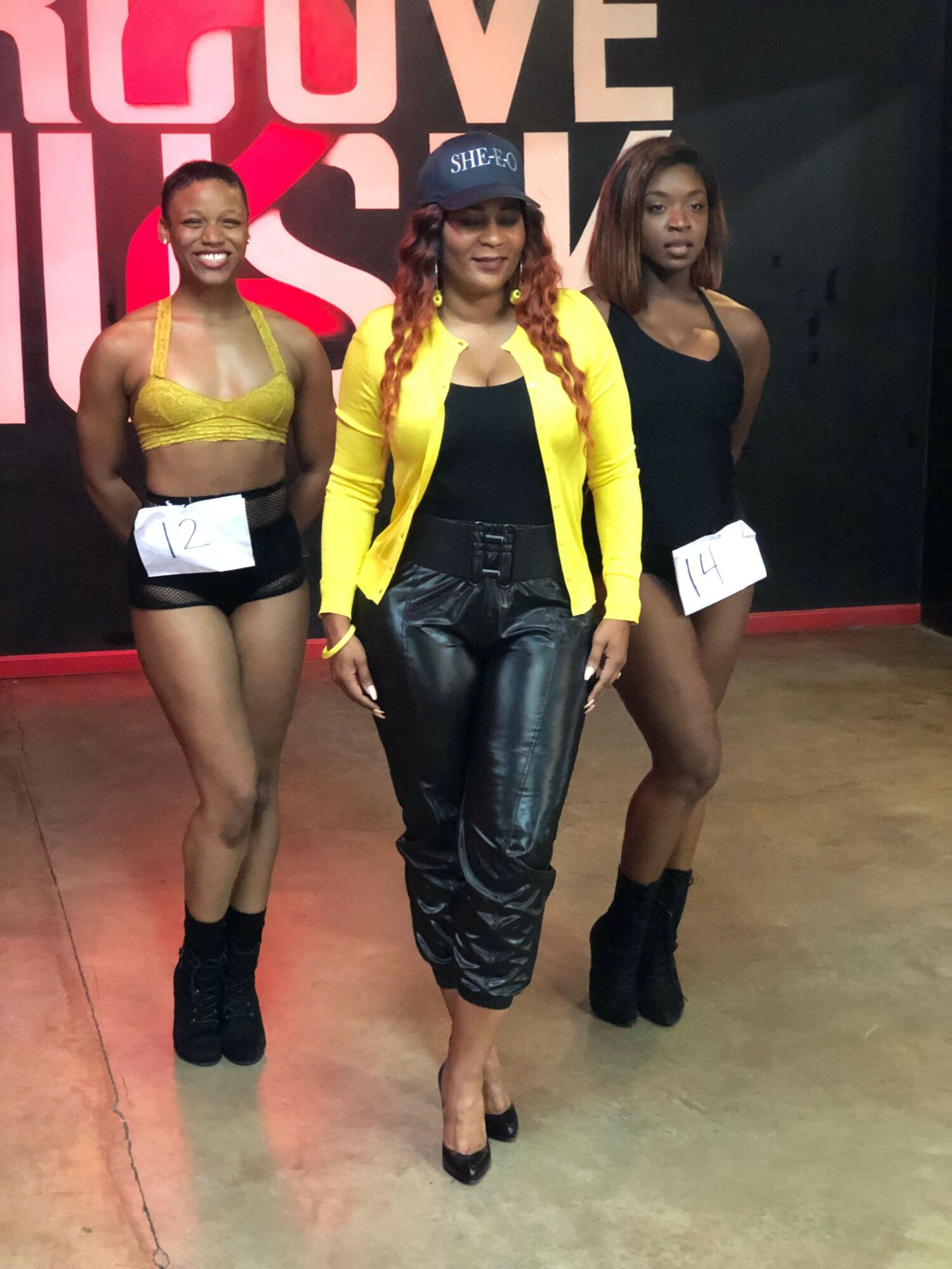 Three women standing together, one in a yellow jacket, black leather pants, and black shoes, in front of a pink and white background.