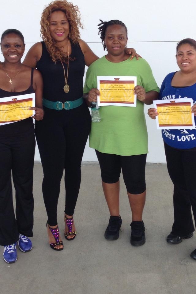 Four women standing outdoors, three holding certificates, smiling, with a white wall and power lines in background.