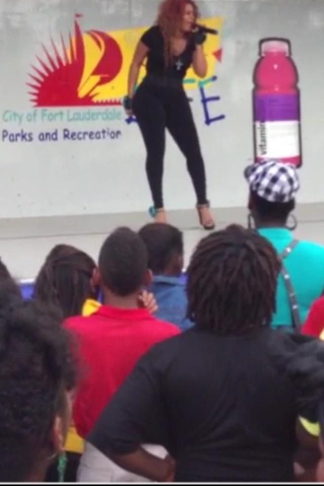 Performer singing on stage with audience watching, colorful banner in background, large water bottle prop, and a person with checkered hat.