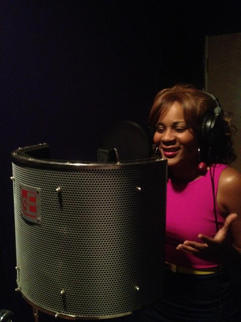 Woman in pink top wearing headphones singing into a microphone in a recording studio.