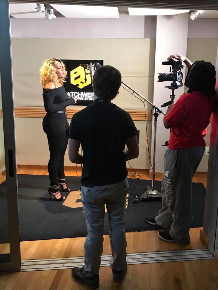 Woman with curly blonde hair being interviewed in a studio, with a camera operator and a person in the foreground, studio background.