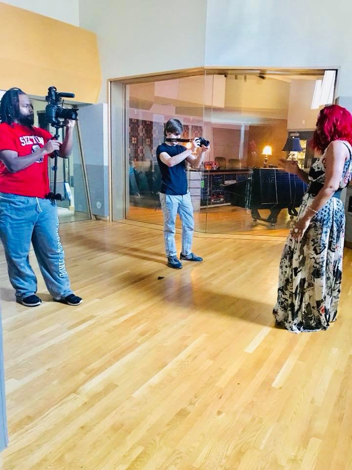 Three people in a recording studio, one with a camera, one with a microphone, and a woman with red hair and a patterned skirt, standing on a wooden floor.