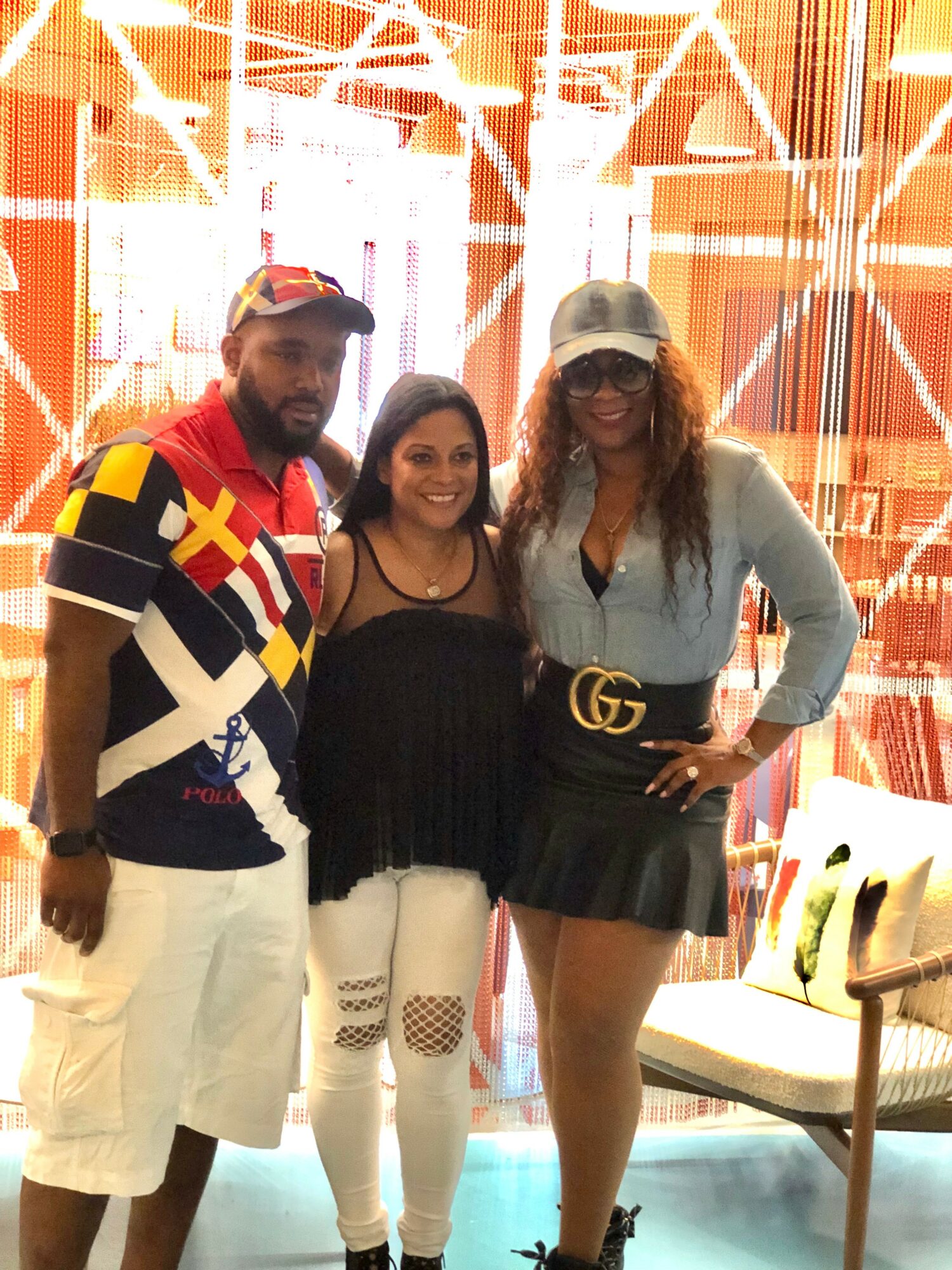 Three people standing indoors with orange and white background, smiling, two women and one man, casual clothing, one woman with sunglasses.