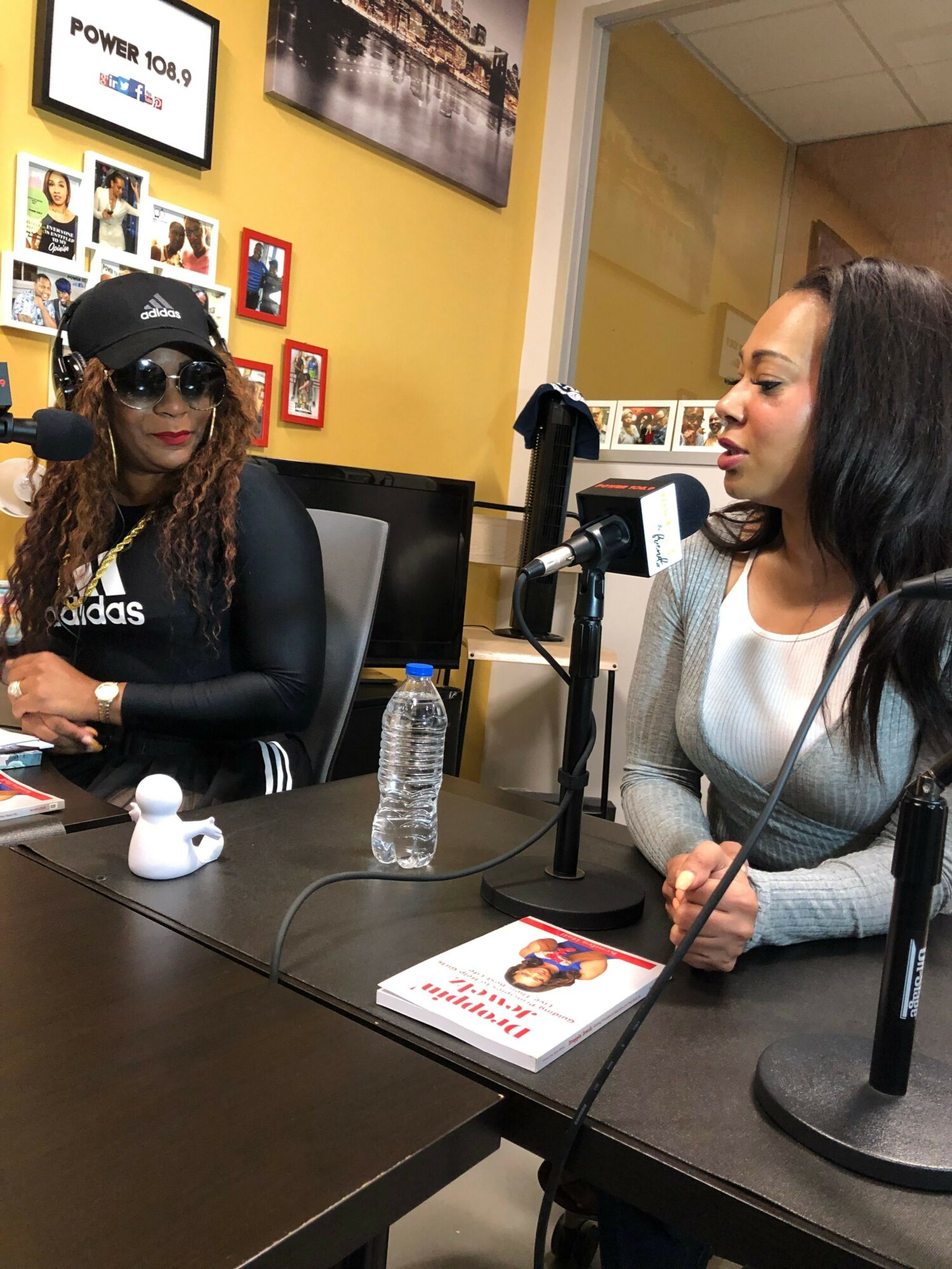 Two women sit at a table with microphones, water bottles, and papers, engaged in a discussion or interview.