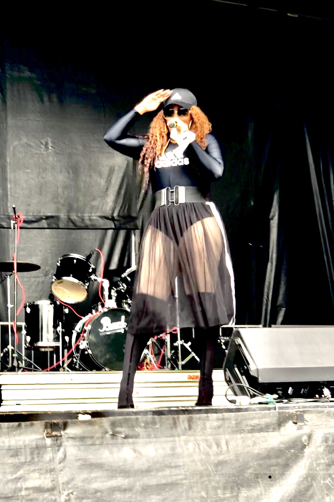 Person on stage wearing a hat, black top, sheer skirt, and high heels, holding a microphone, with musical instruments behind.