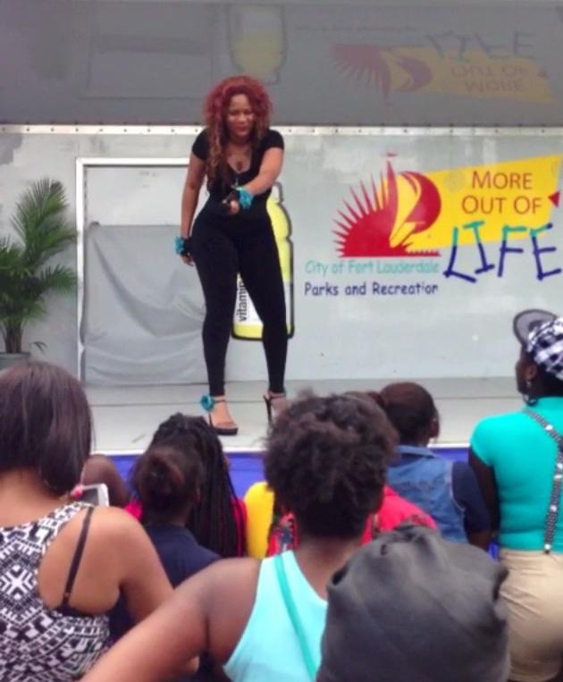 Performer with curly hair on stage, audience watching, outdoor event, banner in background, people seated and standing, sunny day.