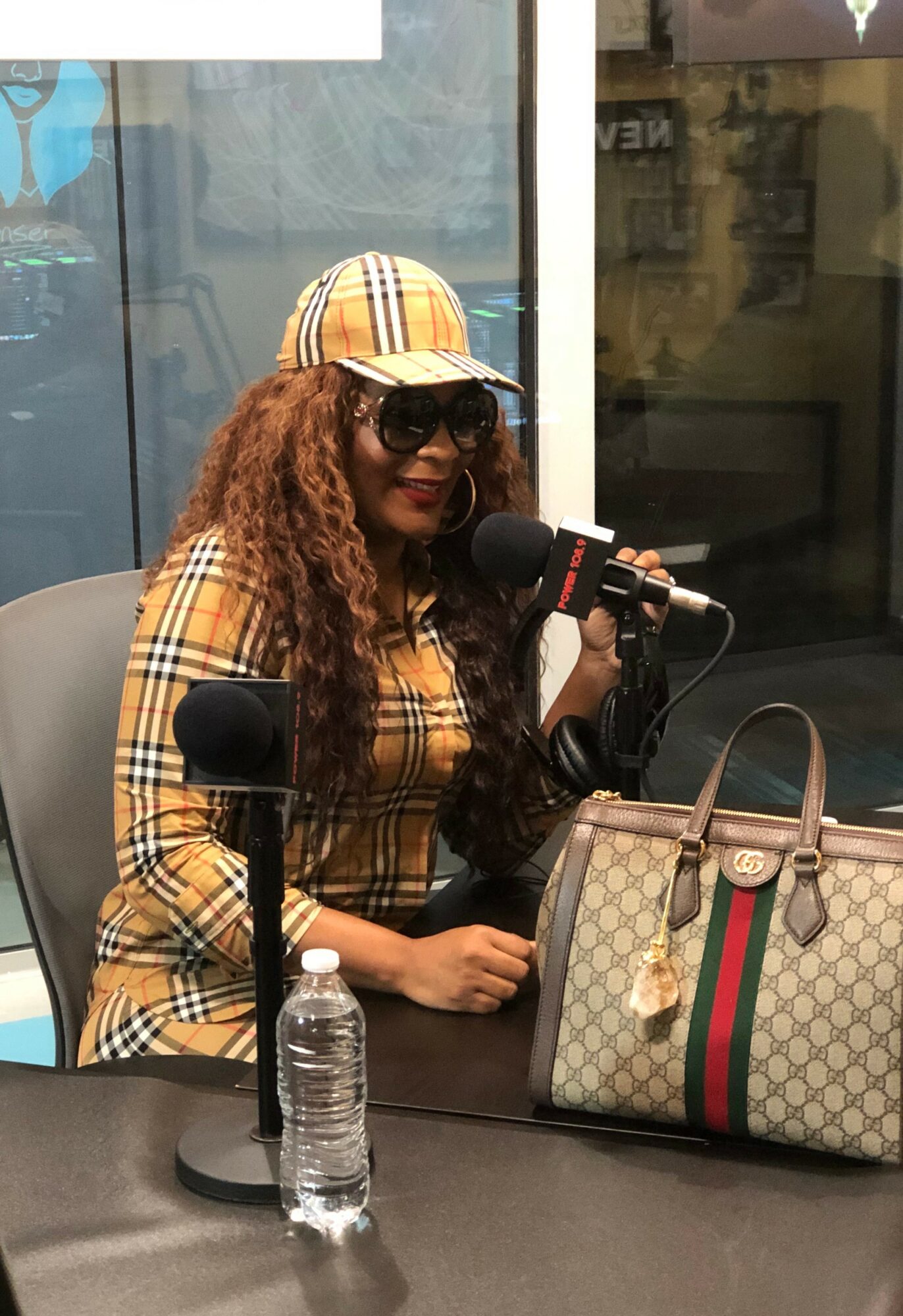 Woman with curly hair wearing a plaid hat and sunglasses, speaking into a microphone, sitting at a table with a patterned handbag and water bottle.