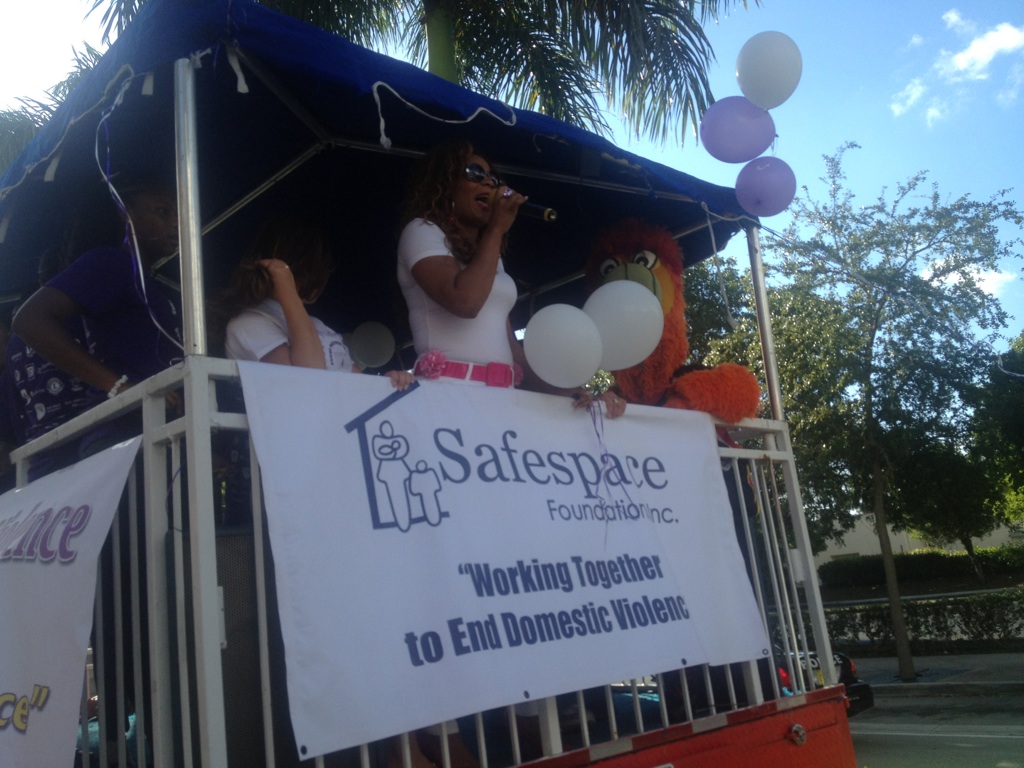 Person speaking into microphone on a decorated outdoor stage with a banner about ending domestic violence, balloons, and a large plush toy.