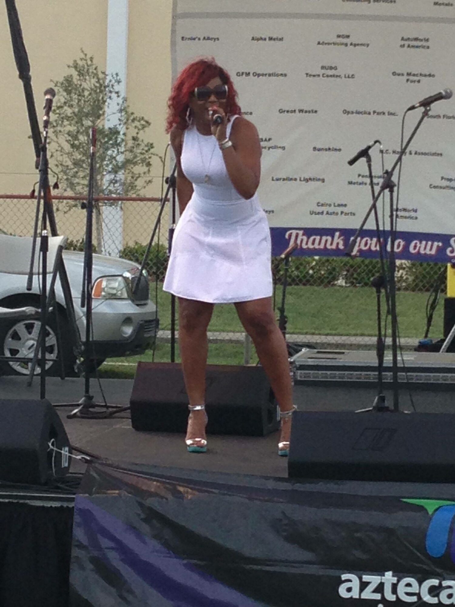 Woman singing on stage with microphone, wearing white dress and sunglasses, outdoor event with trees and cars in background.