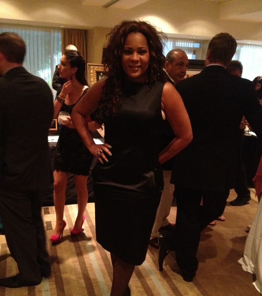 Woman in black dress standing with hands on hips at a social event, surrounded by people in formal attire.