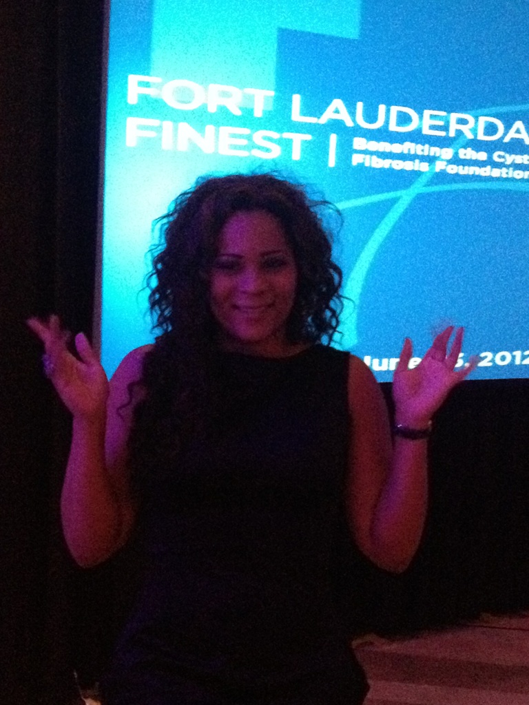 Woman with curly hair smiling and raising hands in front of a blue screen with text.