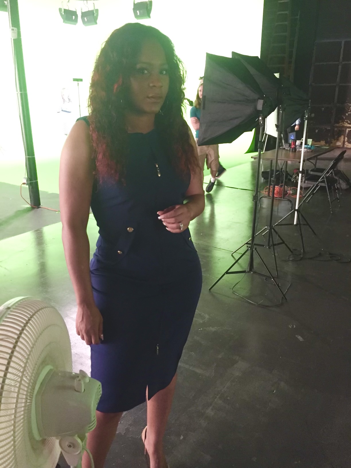 Woman with curly hair standing indoors near lighting equipment, wearing a dark sleeveless top and pants, with a green-lit background.