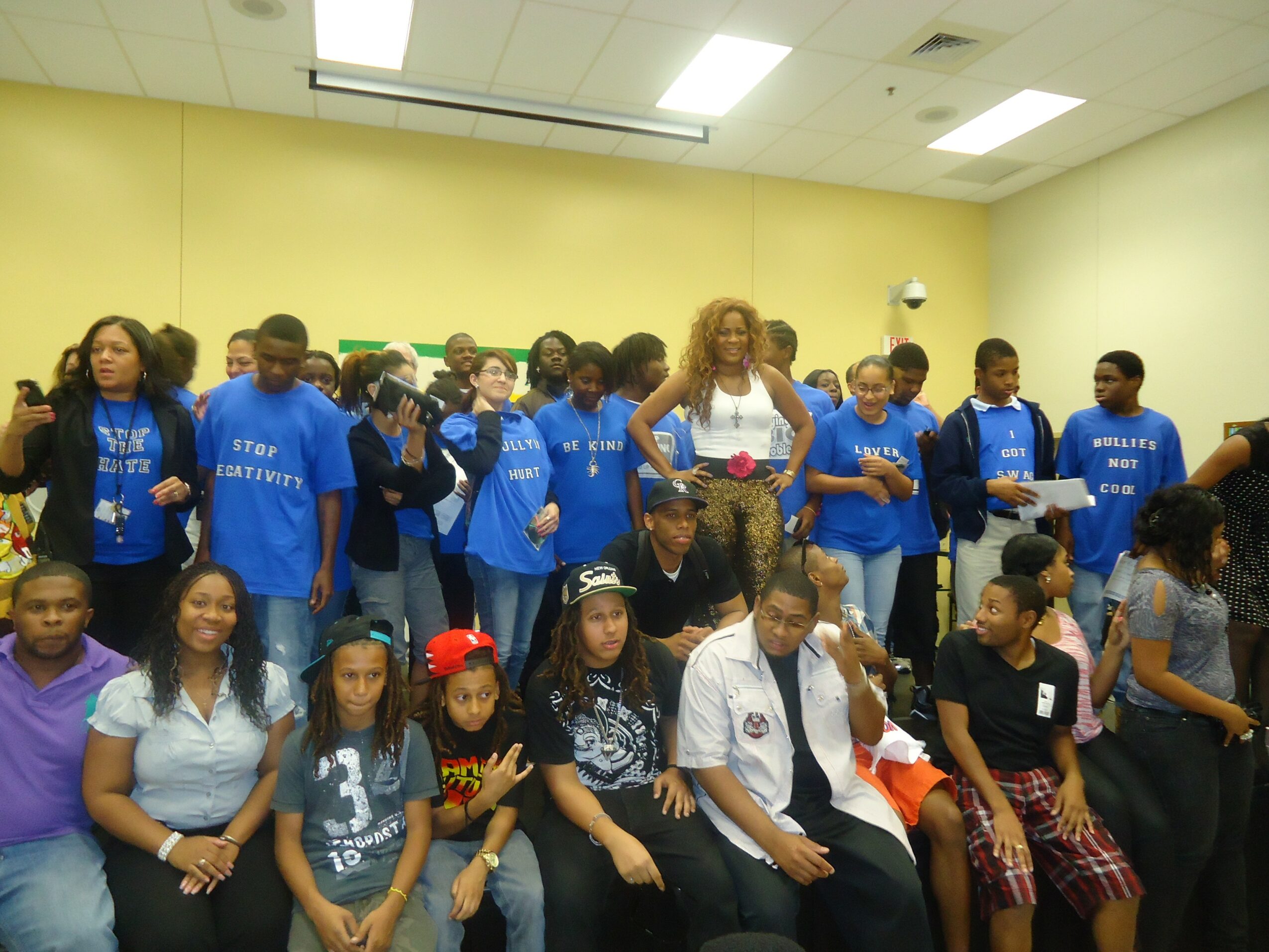 Group of students and a teacher in a classroom, some wearing blue shirts, standing and sitting, with yellow walls.