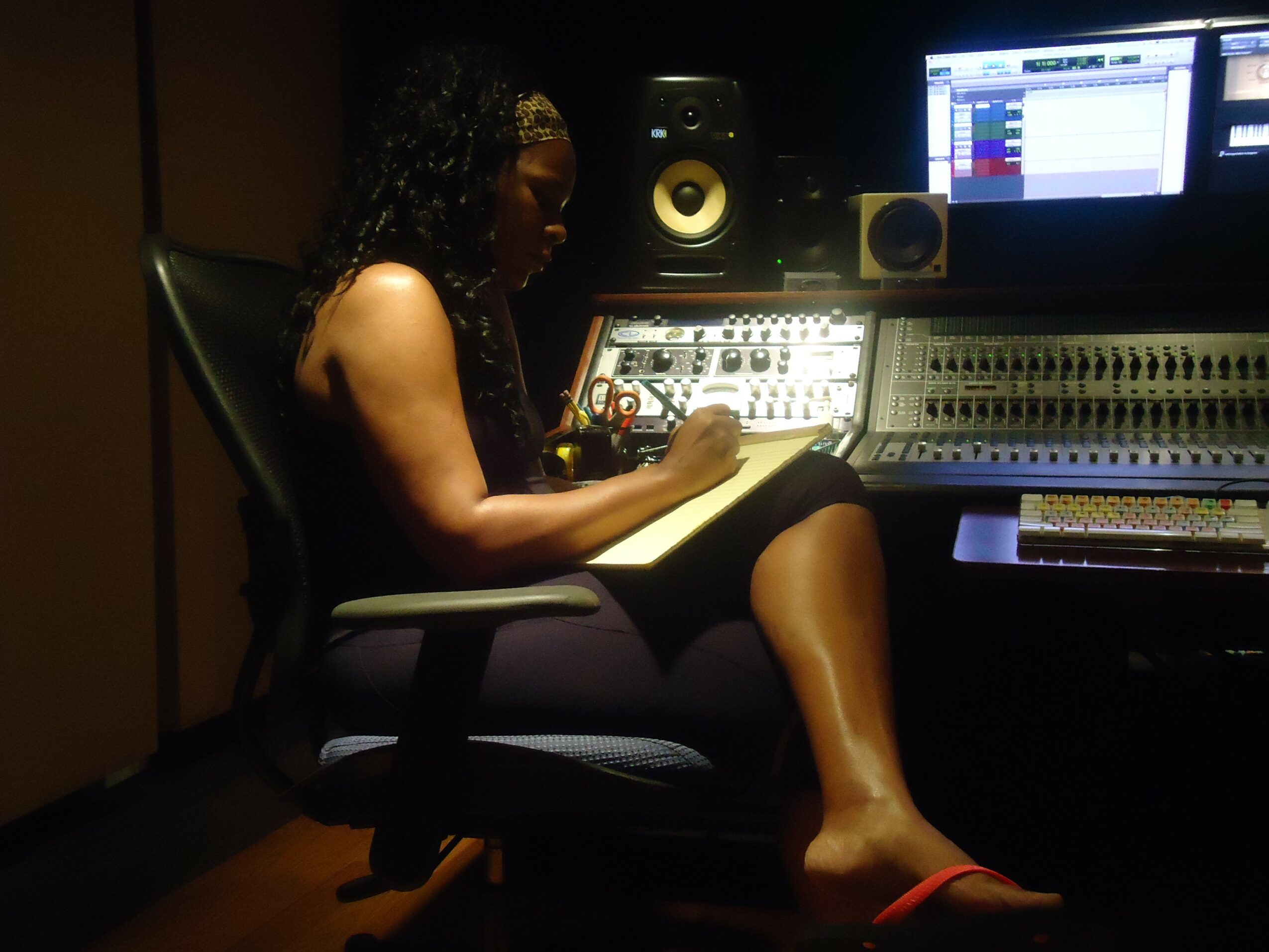 Person sitting in a recording studio, working with audio equipment and a computer screen, in a dimly lit room.