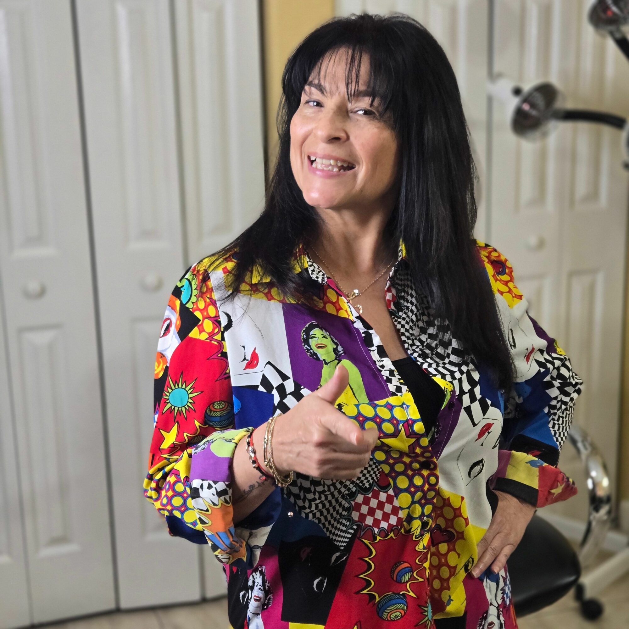 Woman with long dark hair smiling, wearing a colorful patterned jacket, standing indoors in front of white paneled wall.