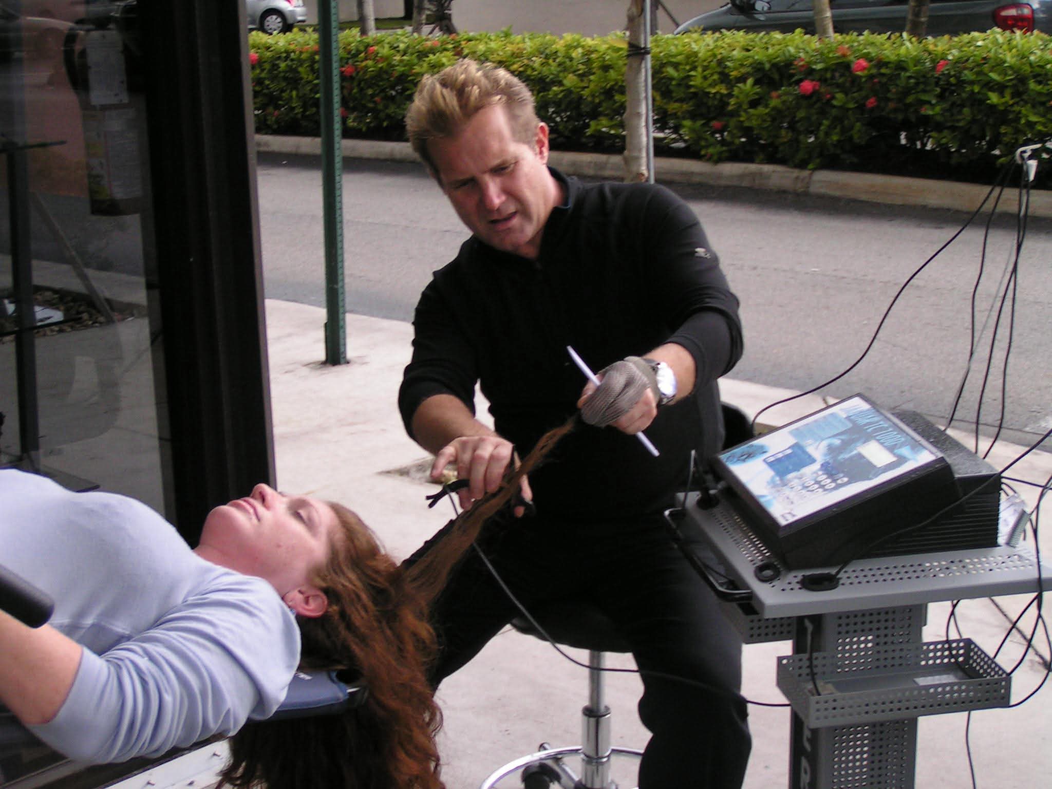 Man performing a medical procedure on a woman lying down outdoors, with equipment nearby, on a sidewalk with bushes and trees.