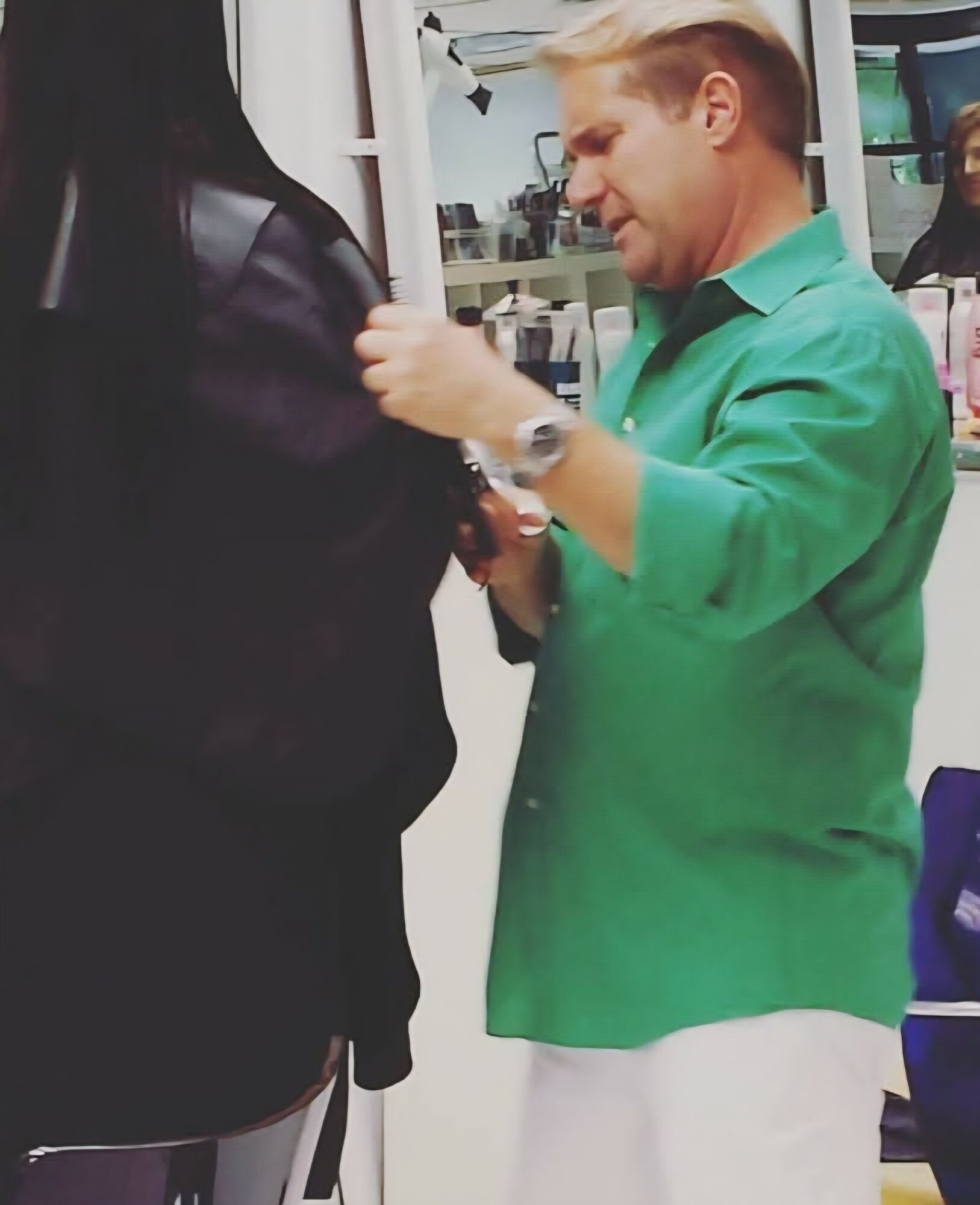 A man in a green shirt and white pants adjusts a woman's black jacket in a store aisle.