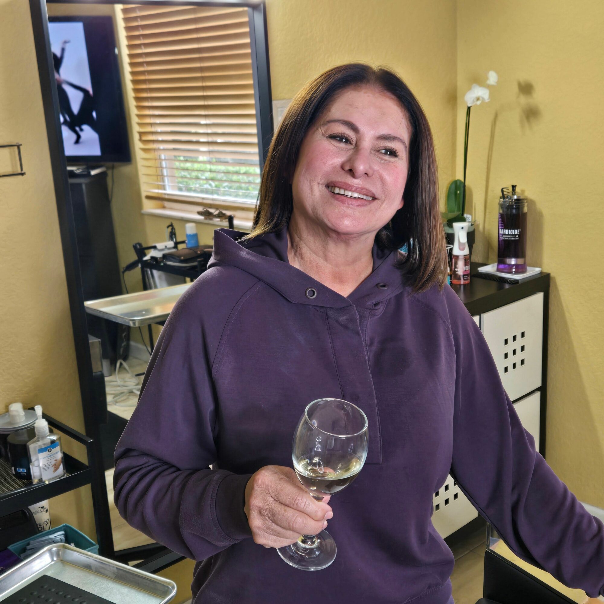 Woman smiling holding a wine glass in a room with a window, bottles, and a computer monitor.