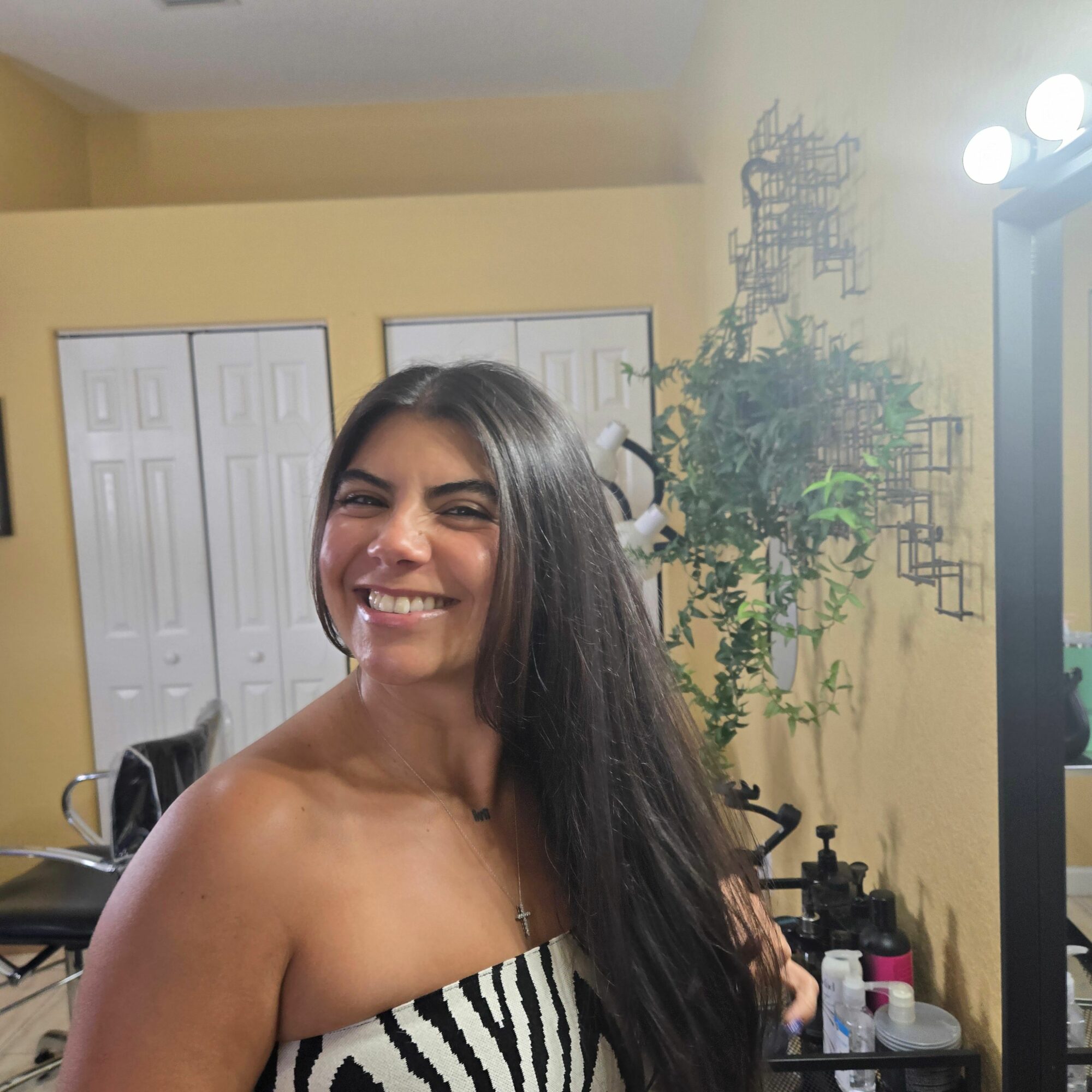 Smiling woman with long dark hair in a striped top, standing indoors with plants and white cabinets in background.