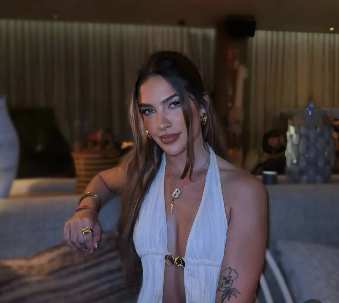 Woman with long hair and tattoos, wearing a white top, sitting indoors with a blurred background.