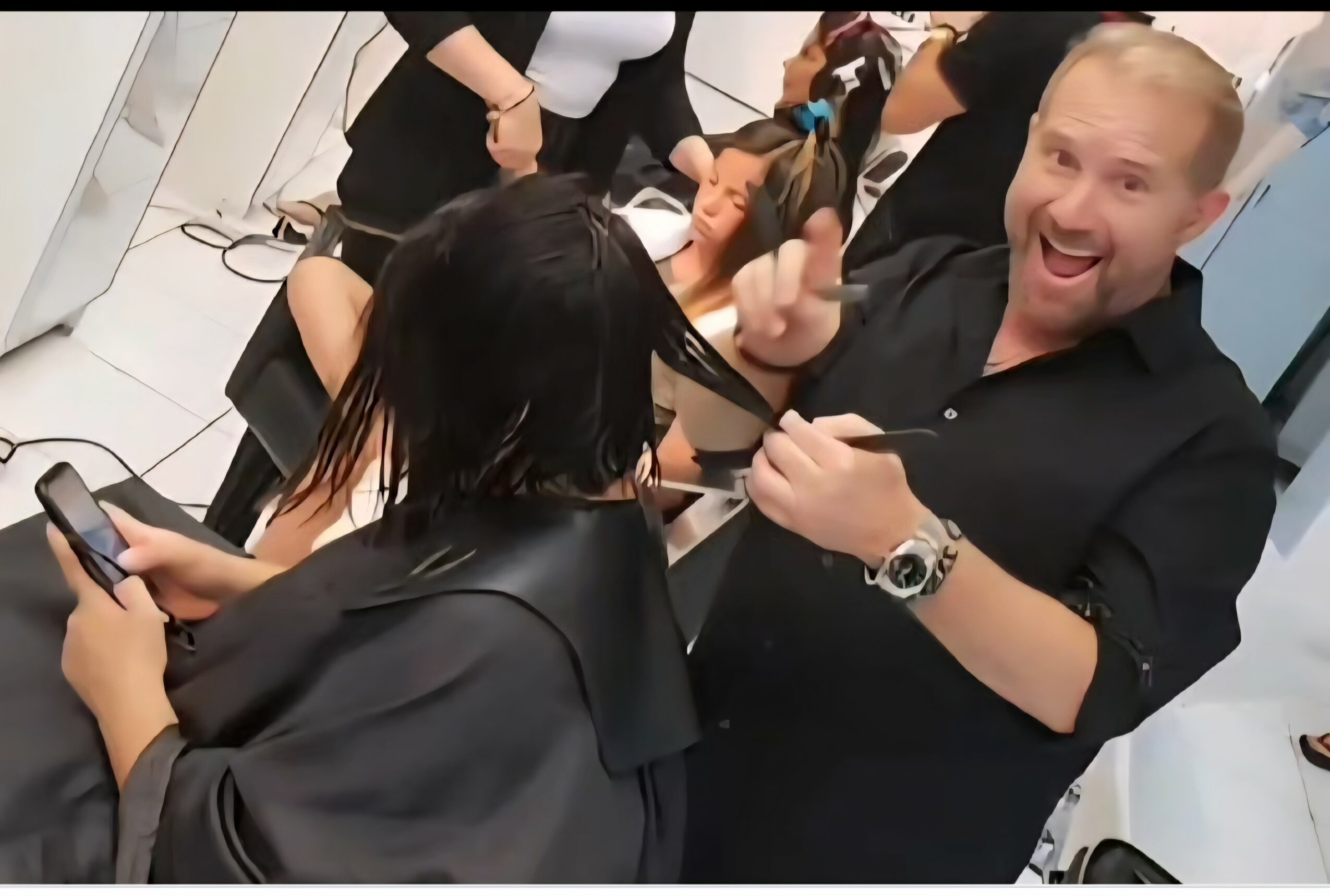 A man with a beard and black shirt smiling and giving a thumbs-up in a busy salon with clients and staff.