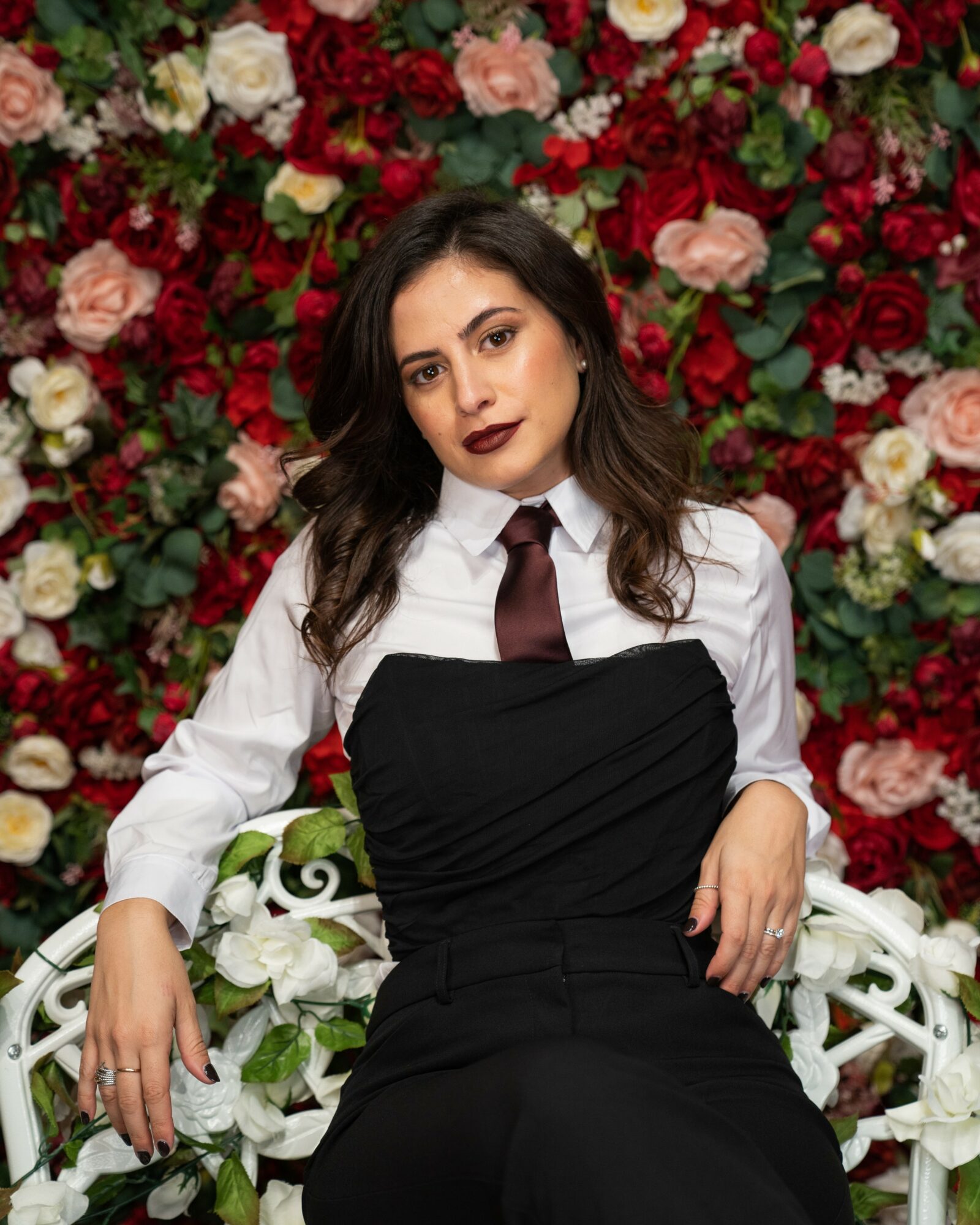 Woman with dark hair and dark lipstick, wearing a white shirt, black dress, and maroon tie, sitting in front of a floral background.