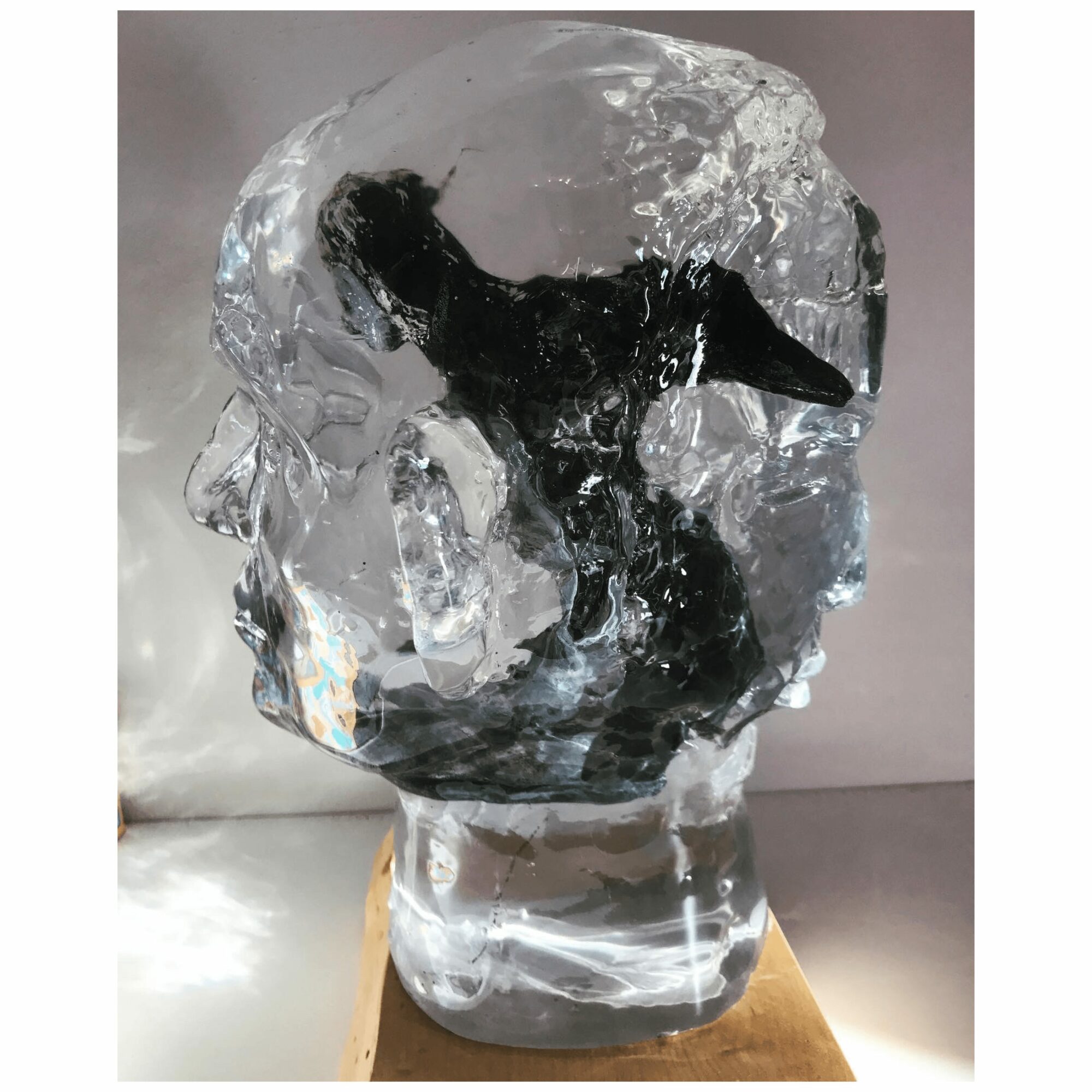 Ice sculpture of a human head with a dark object inside, mounted on a wooden base, against a plain background.