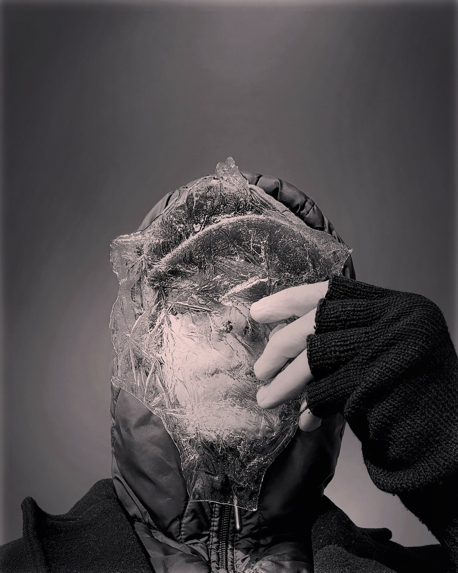 Person wearing a hood and black glove holding a plastic bag over their face, in grayscale.