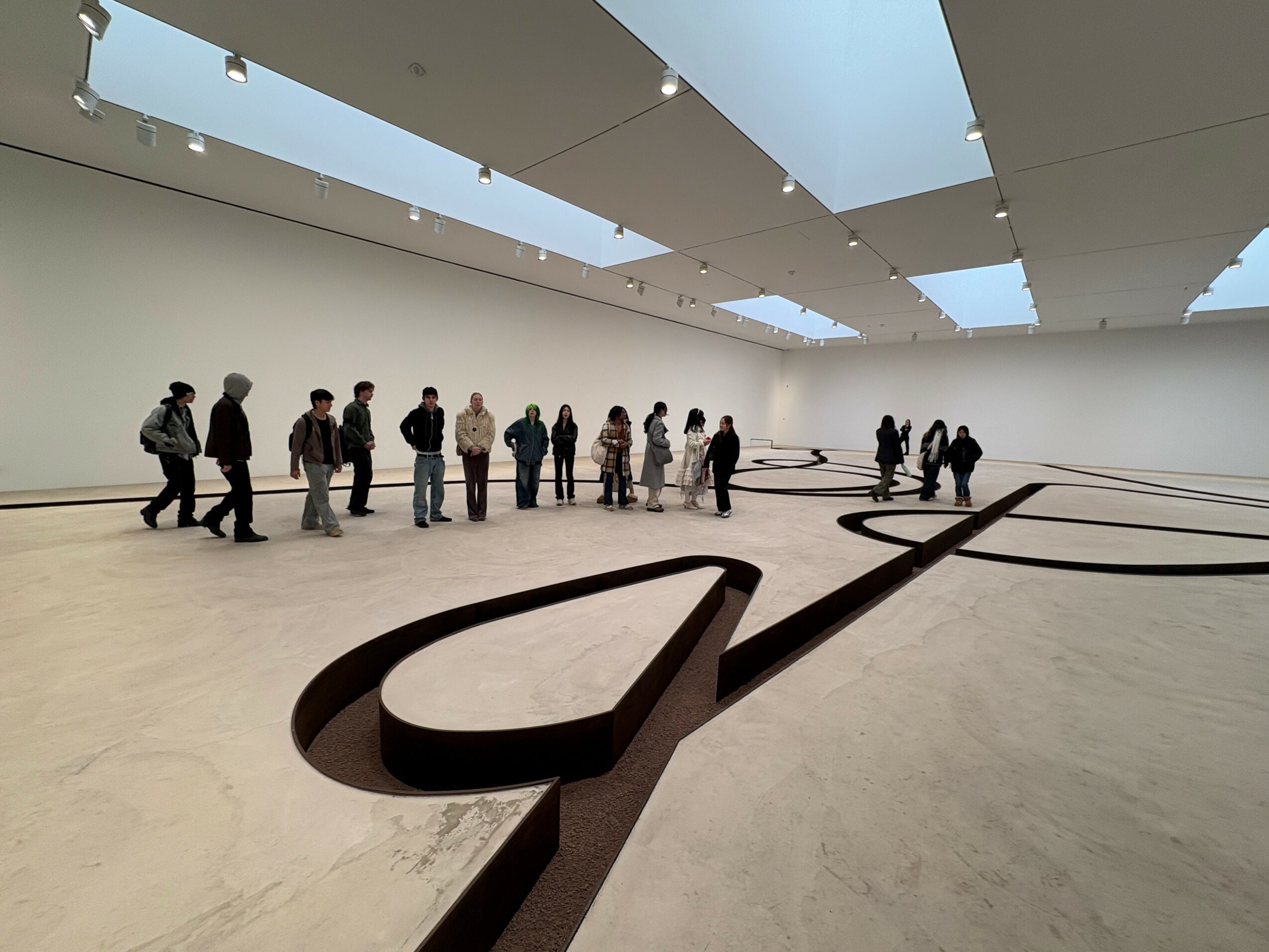 Group of people inside a spacious art gallery with skylights, viewing a large, abstract black sculpture on the floor.