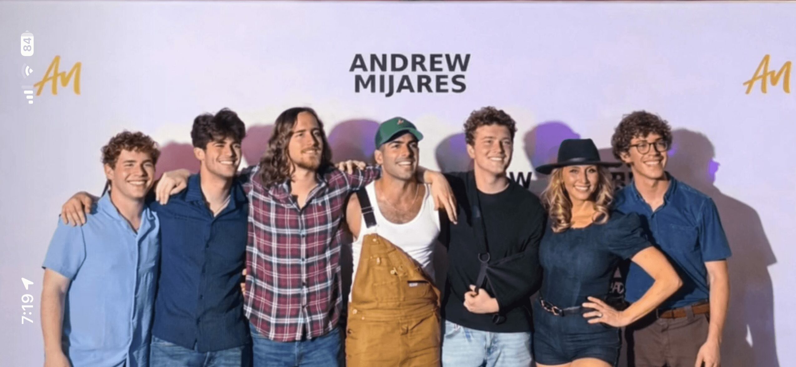 Group of seven people standing together, some smiling, in front of a white background with text. One person is lying down.