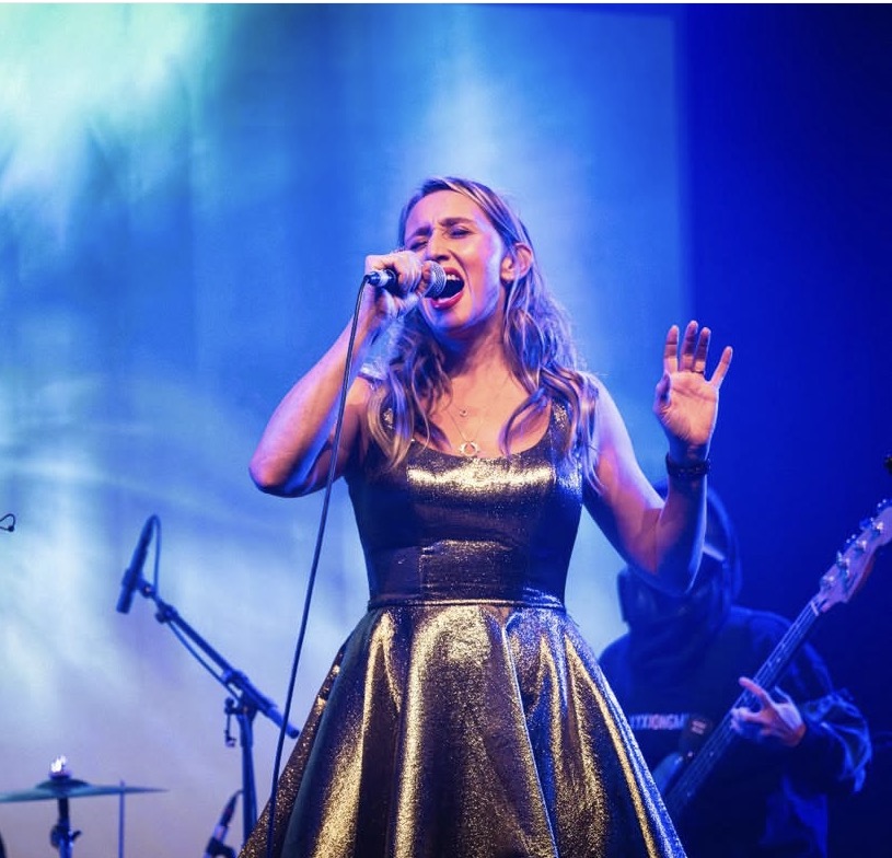 A woman singing into a microphone on stage with blue and green lighting, wearing a shiny dress, with a guitarist in the background.